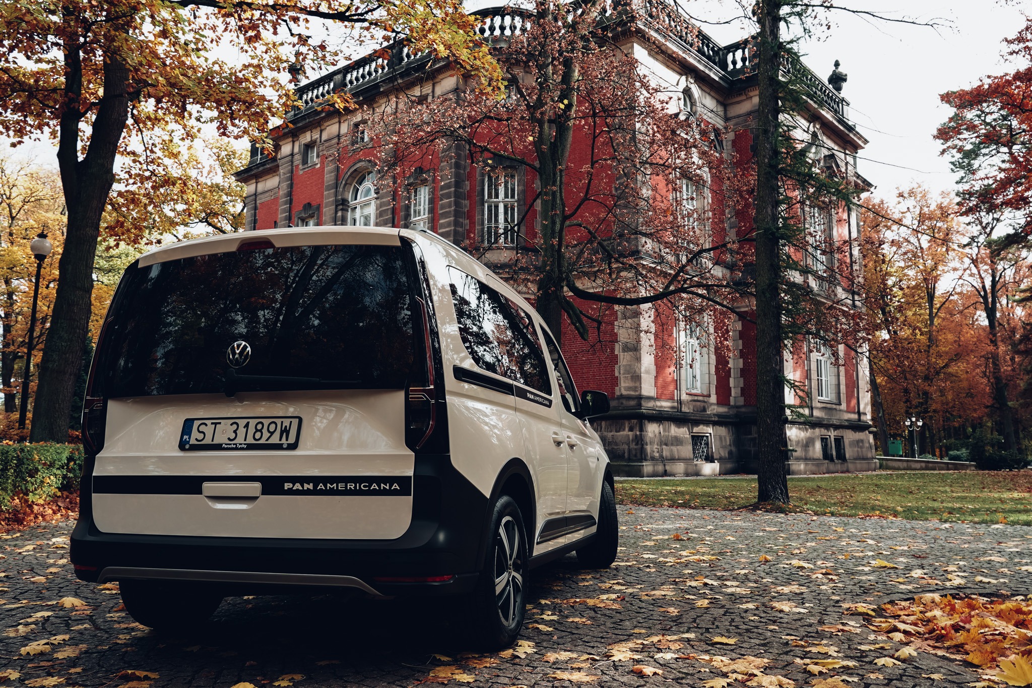Kremowy Volkswagen Caddy PanAmericana na tle ceglanego budynku w jesiennej scenerii. Widok z tyłu, liście na brukowanej drodze.