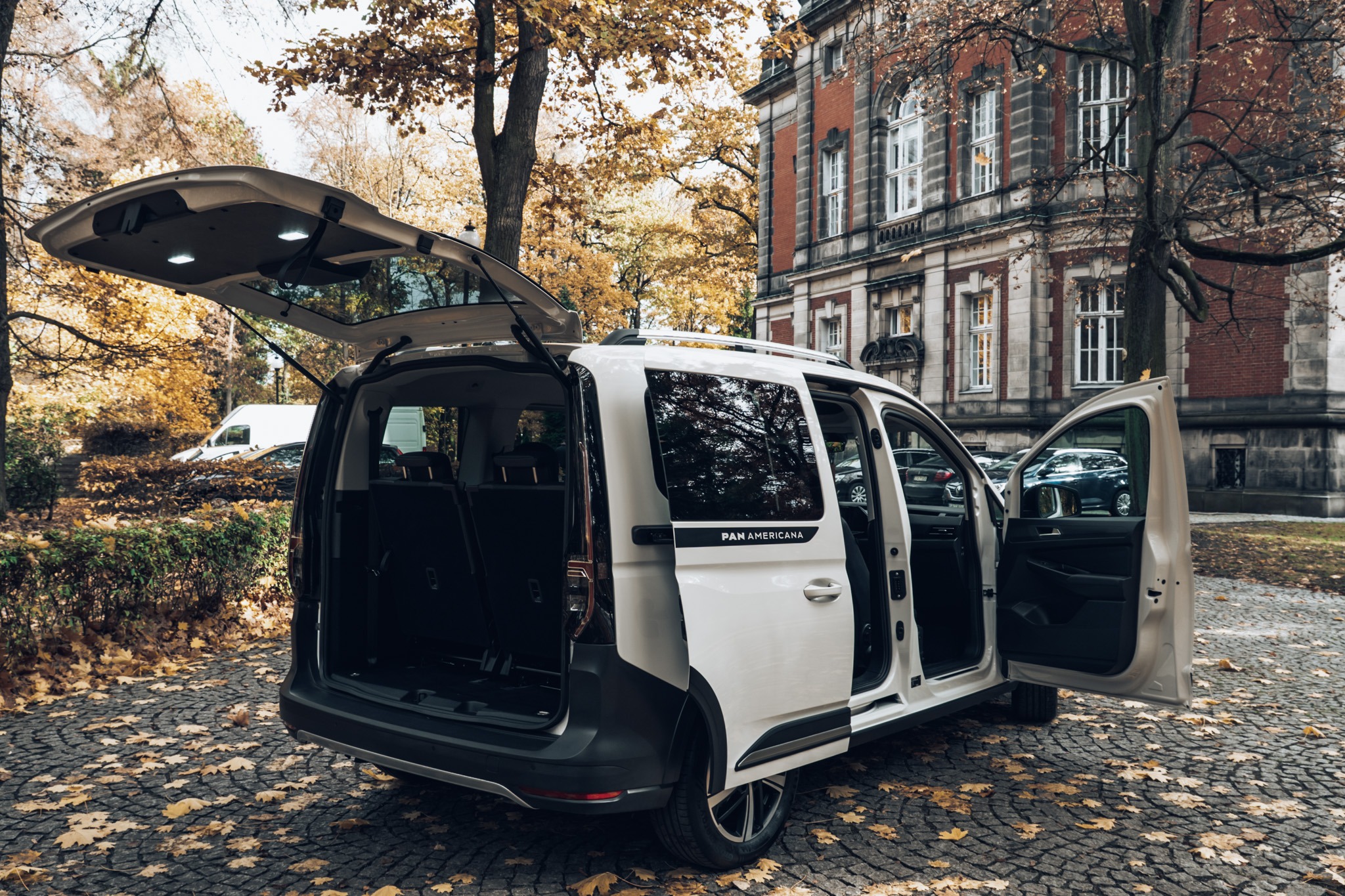 Biały van Pan Americana z otwartymi drzwiami i bagażnikiem na brukowanej drodze, jesienne liście, budynek w tle. Przestronne wnętrze, gotowe do załadunku.