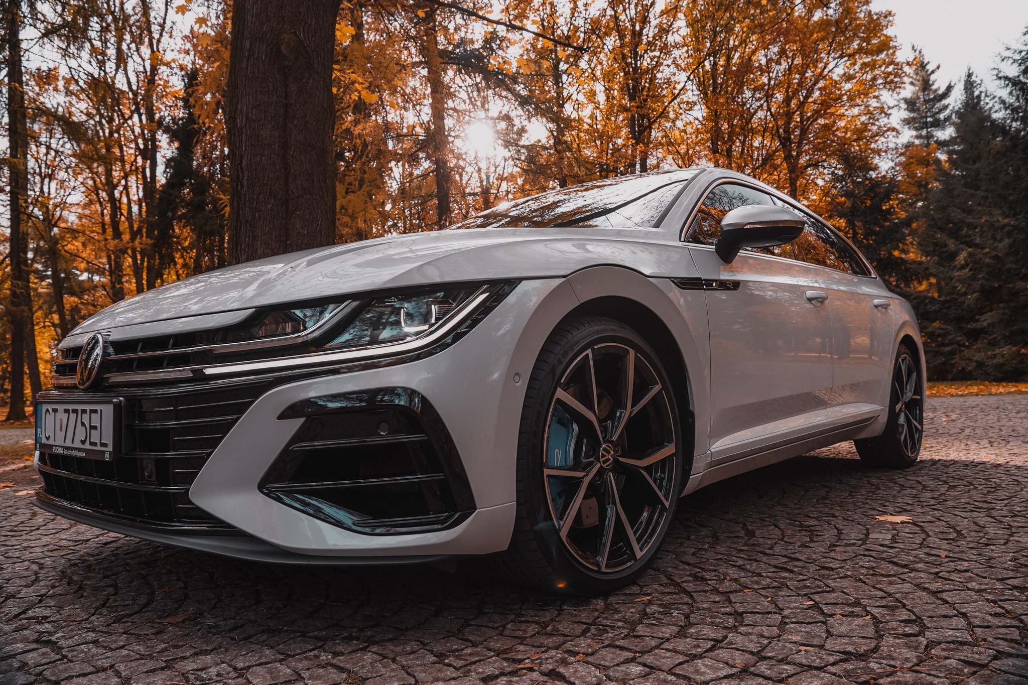 Biały Volkswagen Arteon w jesiennej scenerii parku, z widocznymi detalami felg i reflektorów. Słońce przebija przez korony drzew, oświetlając auto.