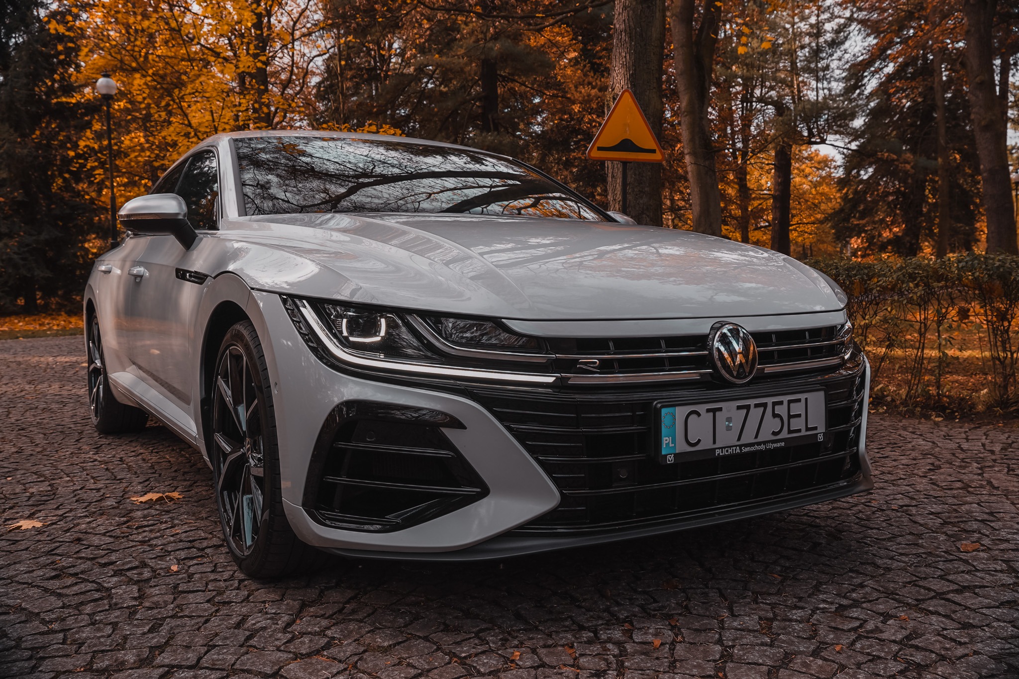 Biały Volkswagen Arteon R na brukowanej drodze, jesienne drzewa w tle, znak drogowy ostrzegający o progu zwalniającym. Detal na przedni grill i reflektory.