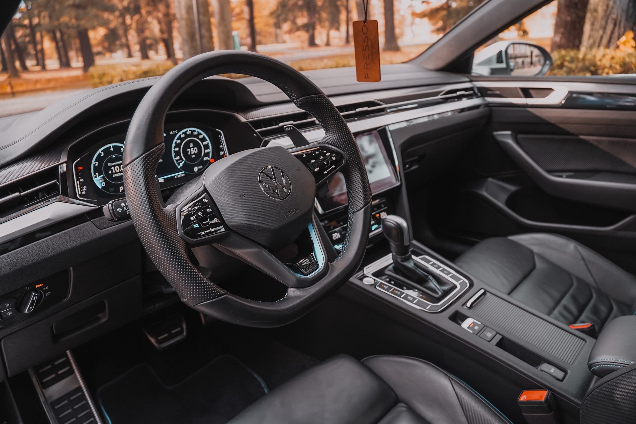 Wnętrze samochodu Volkswagen Arteon z czarną skórzaną tapicerką, widok na deskę rozdzielczą, kierownicę i konsolę środkową. W tle jesienny krajobraz.