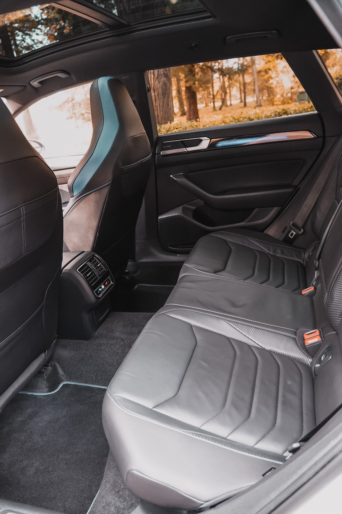 Wnętrze luksusowego samochodu z czarną skórzaną tapicerką, widok na tylne siedzenia z zagłówkami z niebieskimi akcentami i panoramicznym dachem, jesienne drzewa w tle.