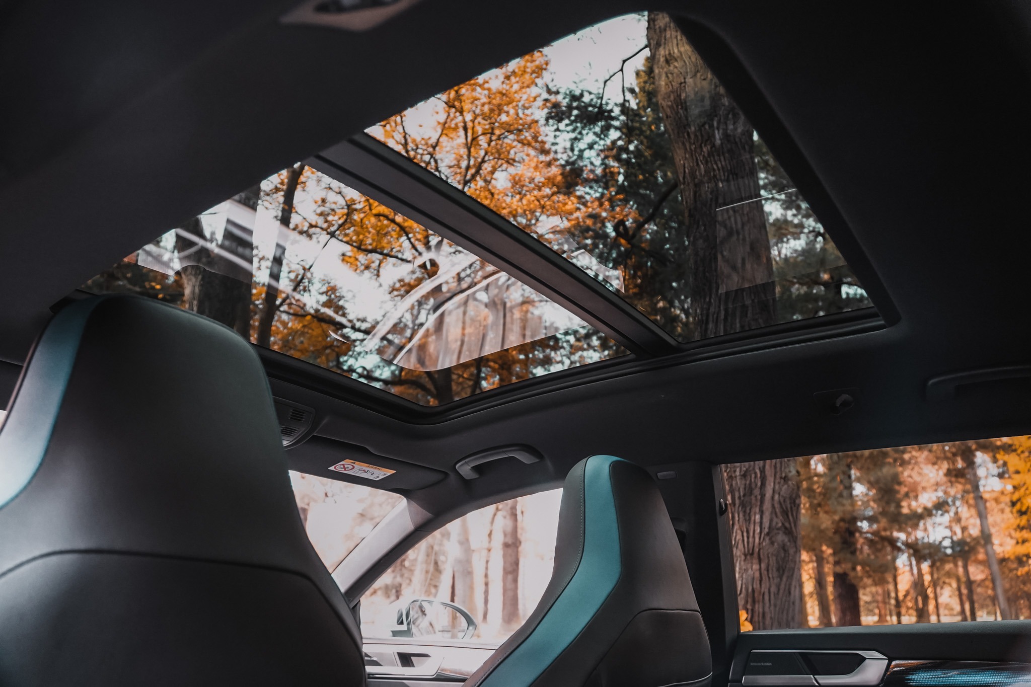 Wnętrze samochodu z widokiem przez panoramiczny dach na jesienny las. Widoczne fotele z niebieskimi akcentami i fragment deski rozdzielczej.