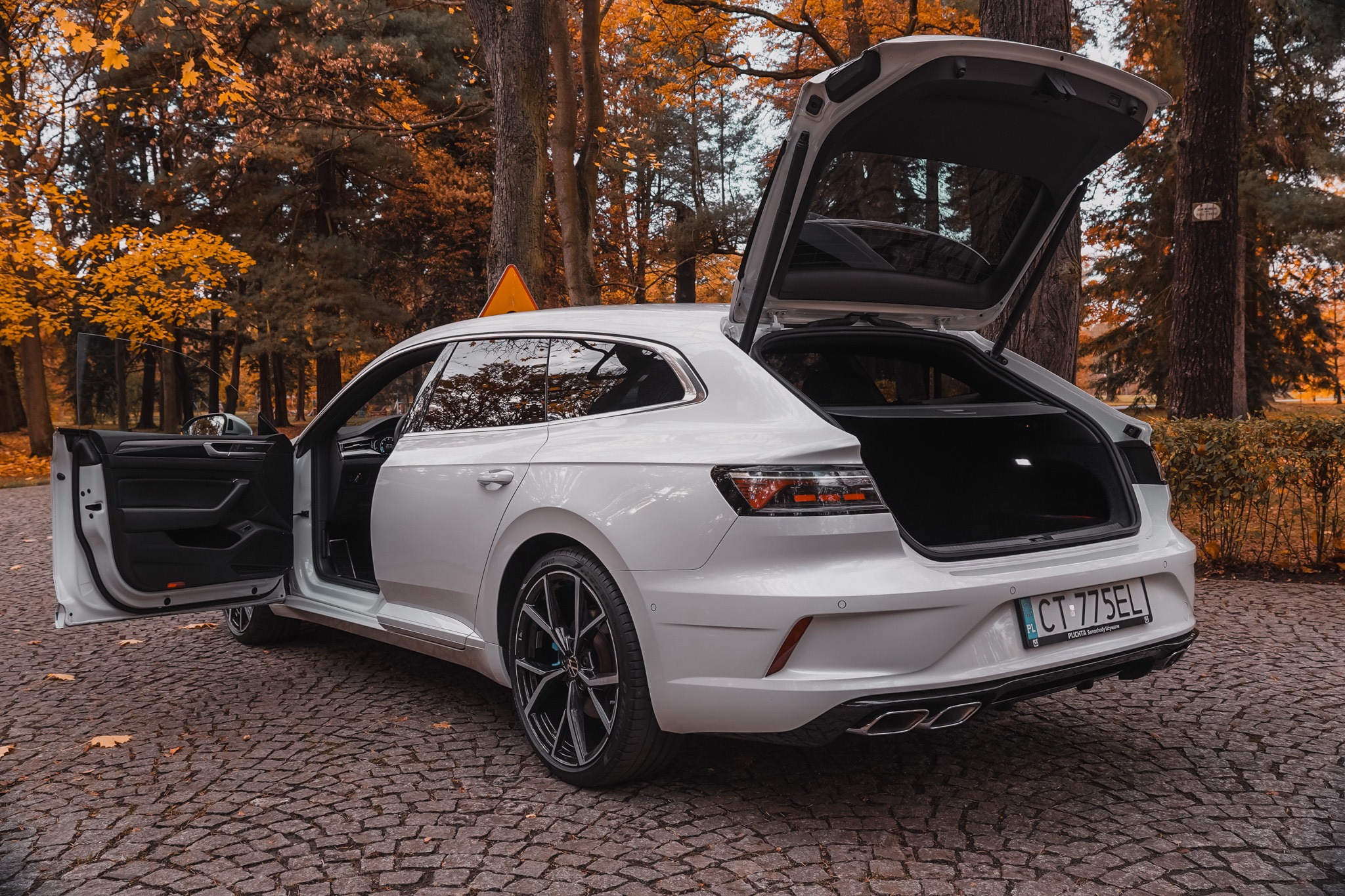 Białe kombi z otwartą klapą bagażnika i drzwiami pasażera, zaparkowane na brukowanej drodze w jesiennym otoczeniu drzew. Widoczna tablica rejestracyjna.
