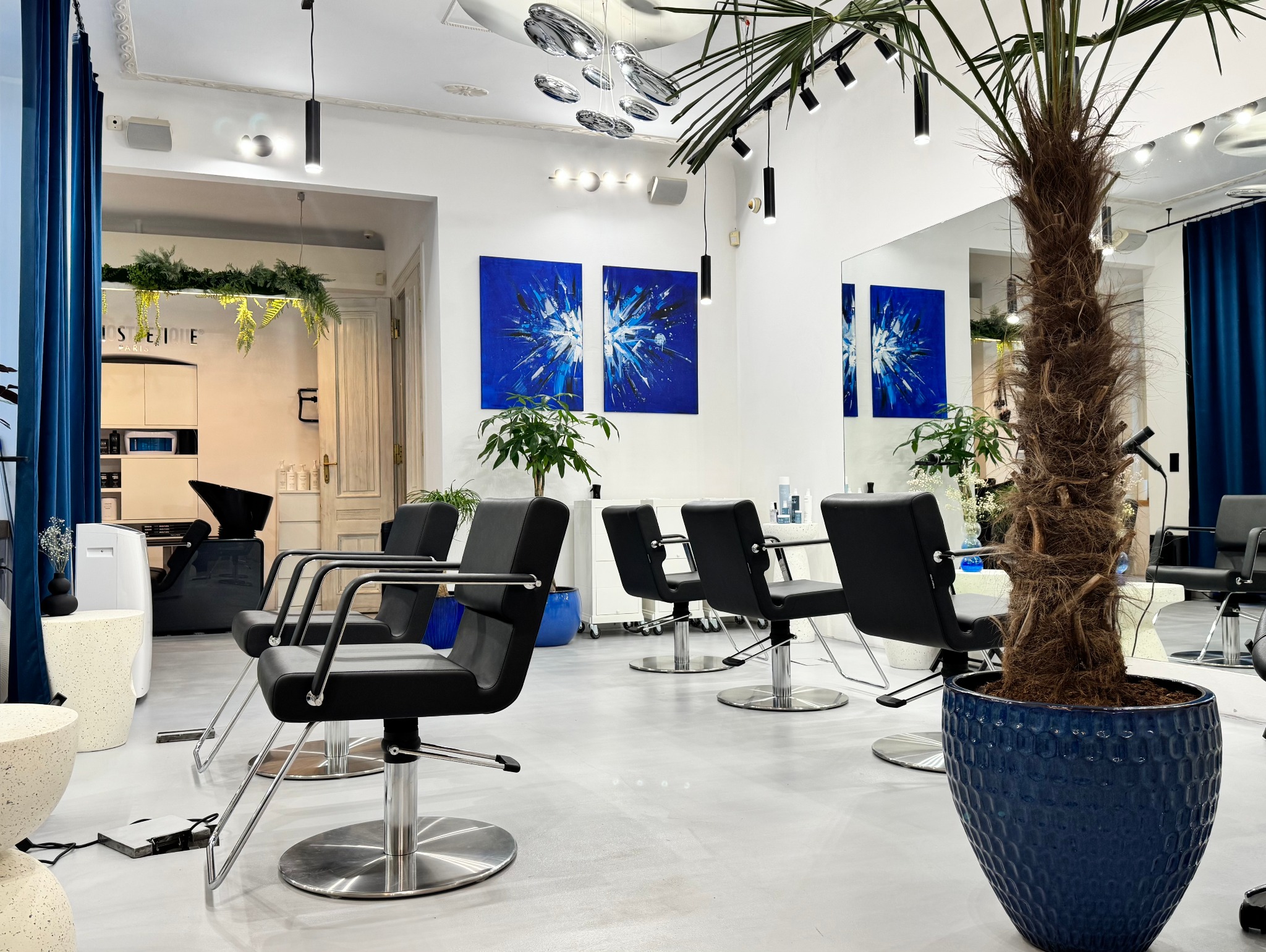 Jasne wnętrze salonu z czarnymi fotelami, lustrami i roślinami w niebieskich donicach. Nowoczesne oświetlenie i dekoracje na ścianach tworzą elegancki klimat.