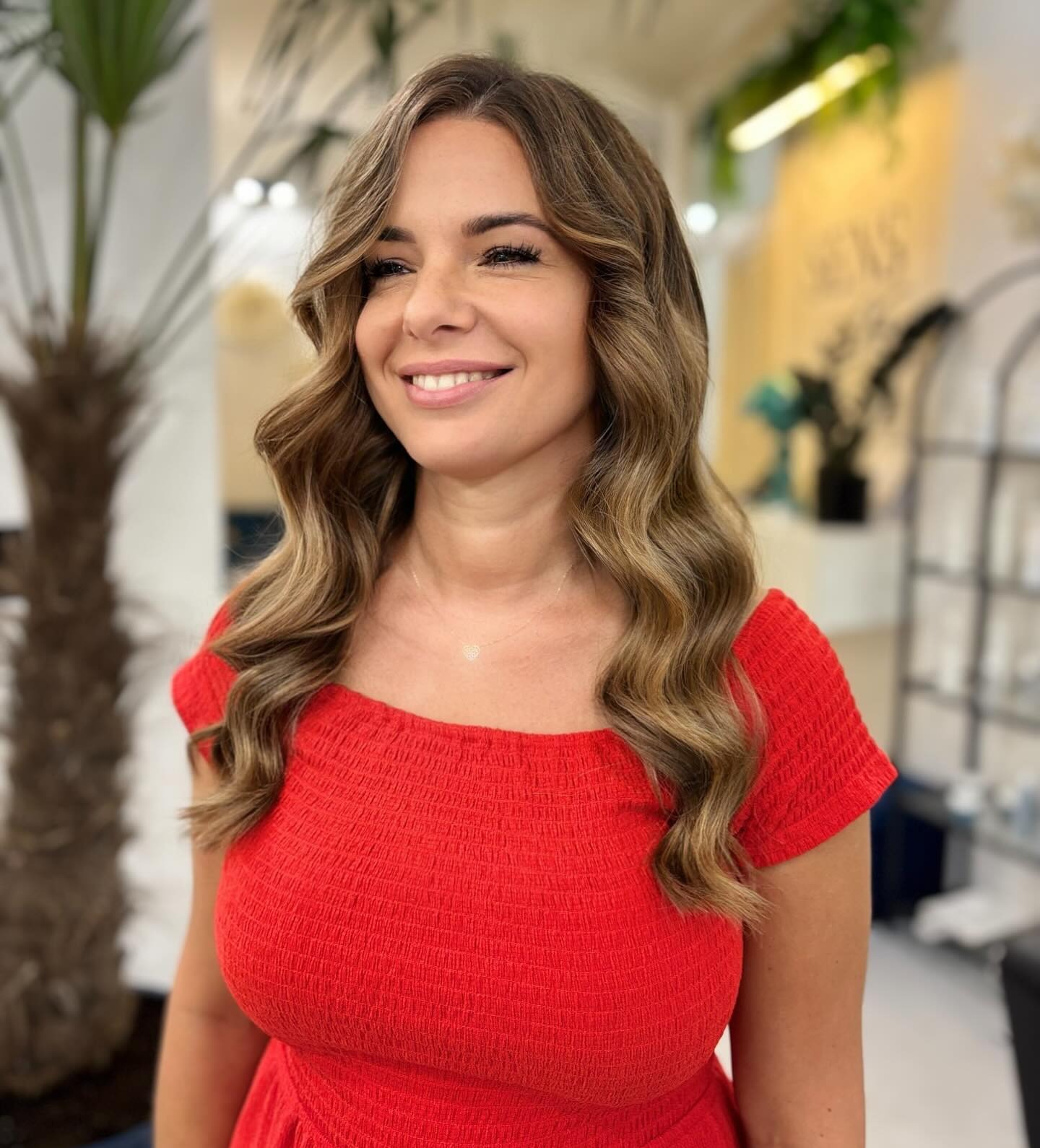 Uśmiechnięta brunetka w czerwonej sukience prezentuje stylizację włosów z delikatnymi falami, wykonaną w salonie fryzjerskim z elementami dekoracyjnymi.