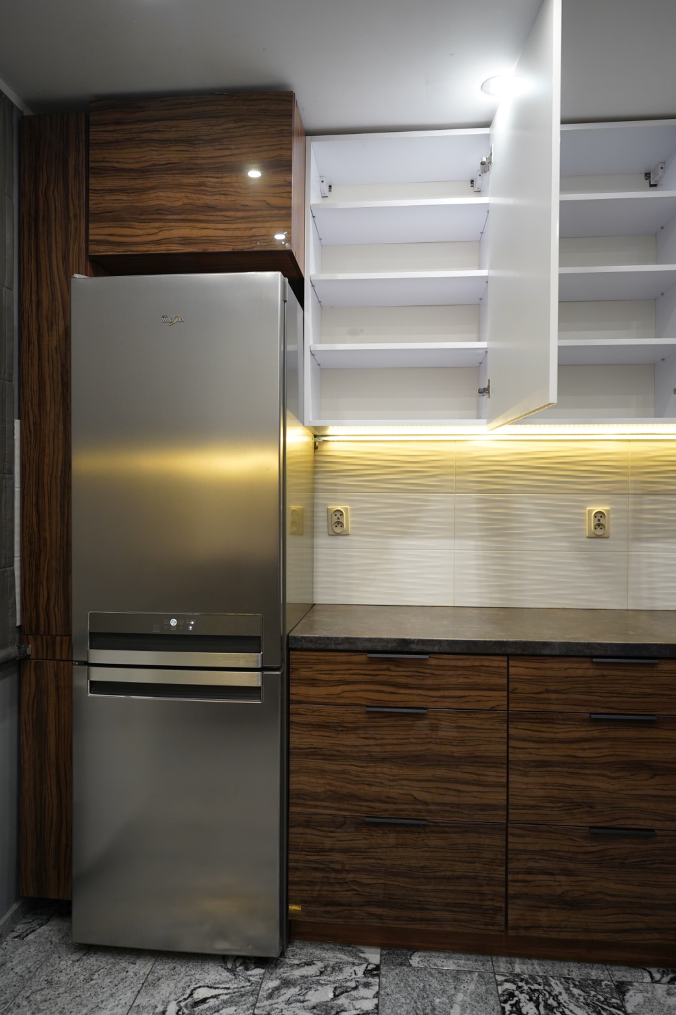 Nowoczesna kuchnia z drewnianymi szafkami o unikalnym wzorze słojów, białymi szafkami wiszącymi i srebrną lodówką Whirlpool. Oświetlenie LED pod szafkami.