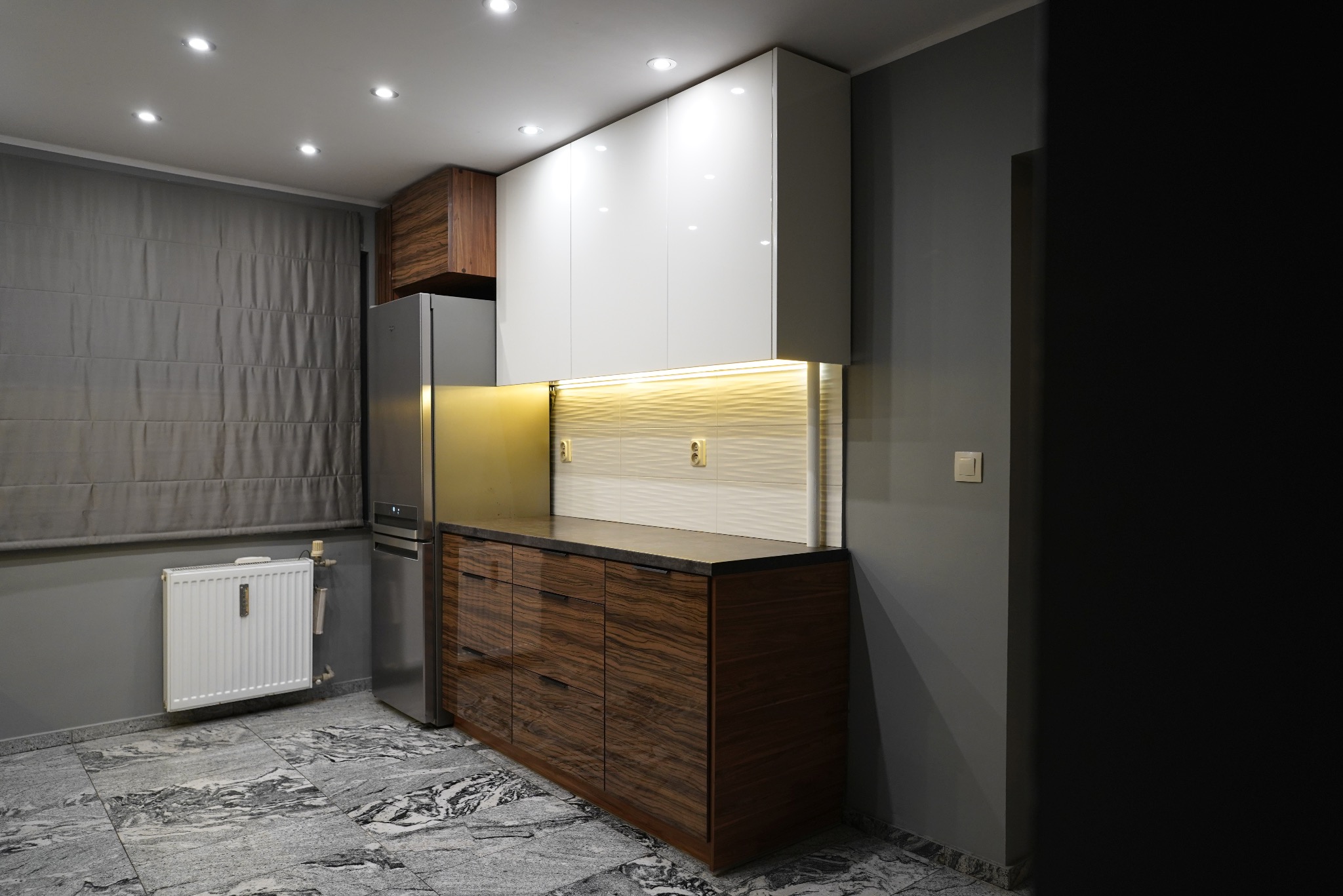 Nowoczesna kuchnia z drewnianymi frontami szafek dolnych i białymi górnymi, podświetlona listwą LED. Na podłodze marmurowe płytki. Widoczny fragment grzejnika.