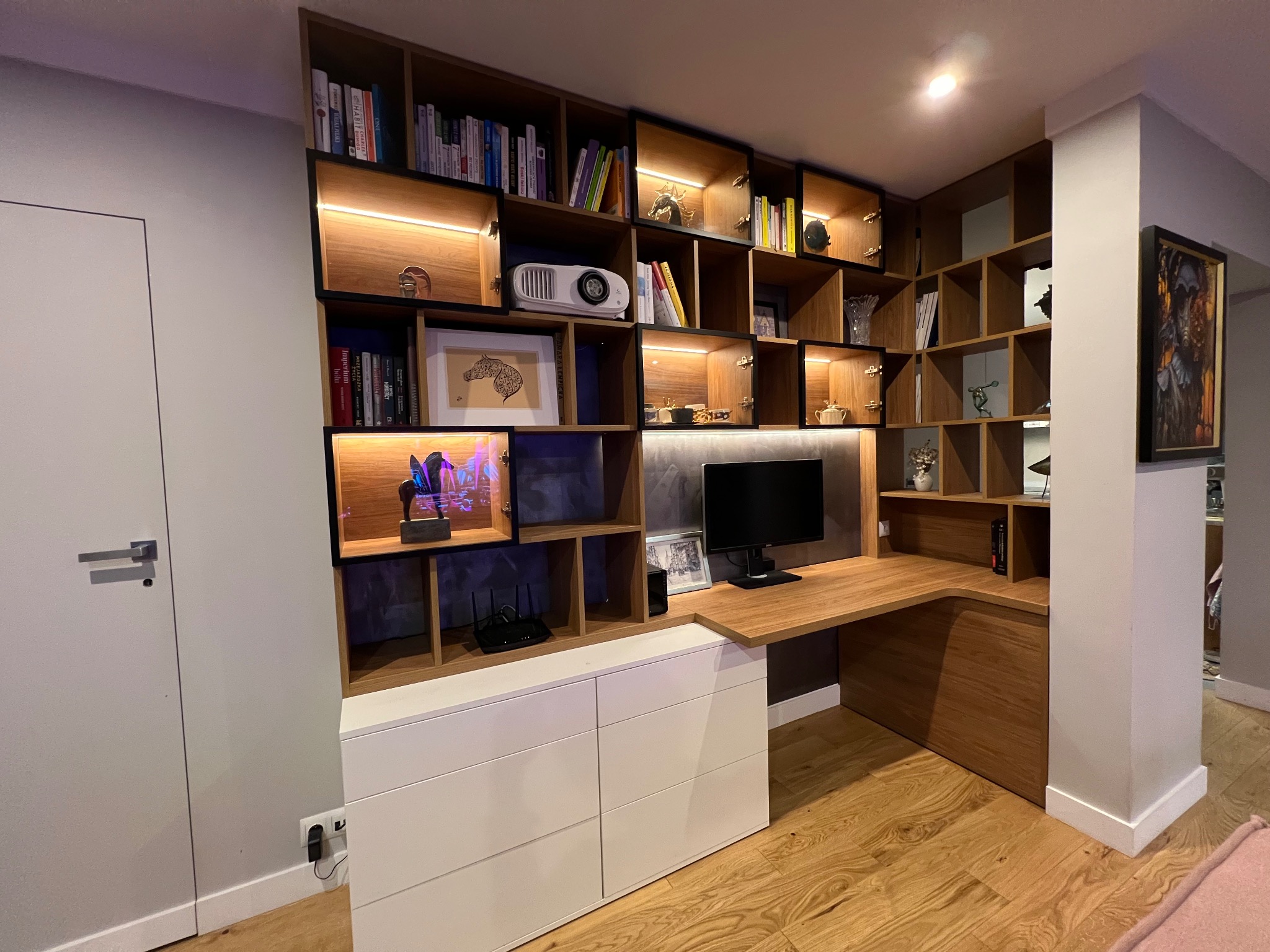 Funkcjonalna zabudowa biurowa z drewnianymi półkami i blatem, oświetleniem LED i białymi szafkami. Na półkach książki, dekoracje i projektor. Nowoczesny design.