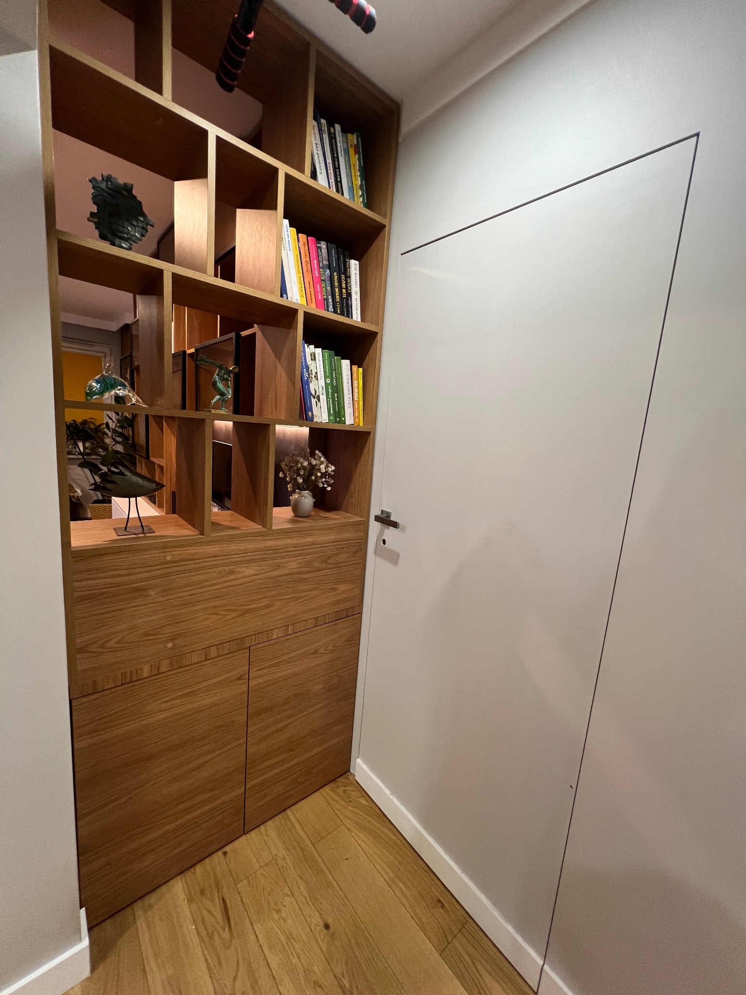Drewniana zabudowa z półkami i szafkami, pełna książek i dekoracji, przylegająca do białej ściany z ukrytymi drzwiami. Ciepłe oświetlenie wnętrza.