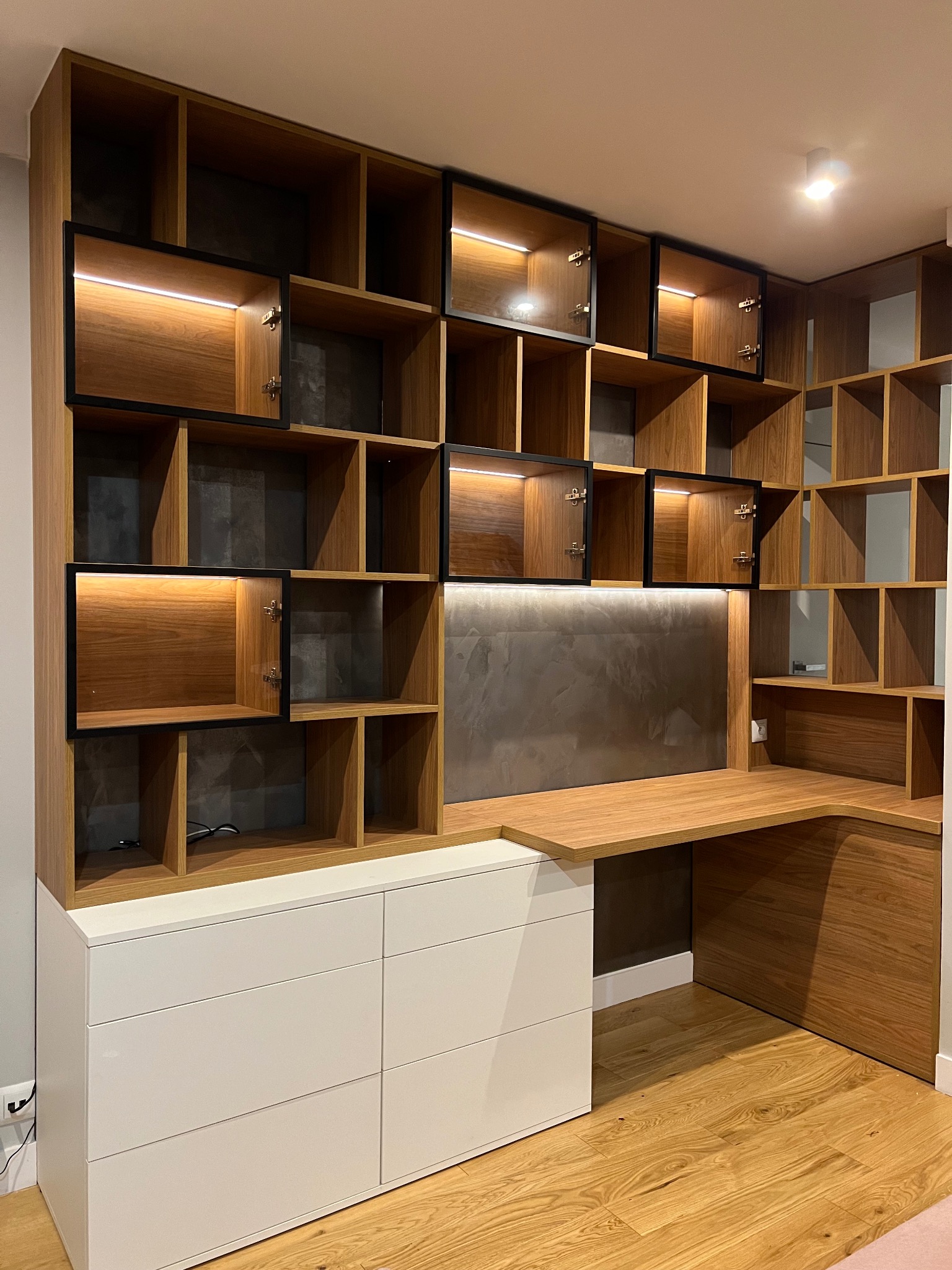 Funkcjonalna zabudowa z biurkiem i szafkami z drewna, z białymi szufladami na dole i oświetleniem LED w górnych szafkach, tworząca nowoczesną przestrzeń do pracy.