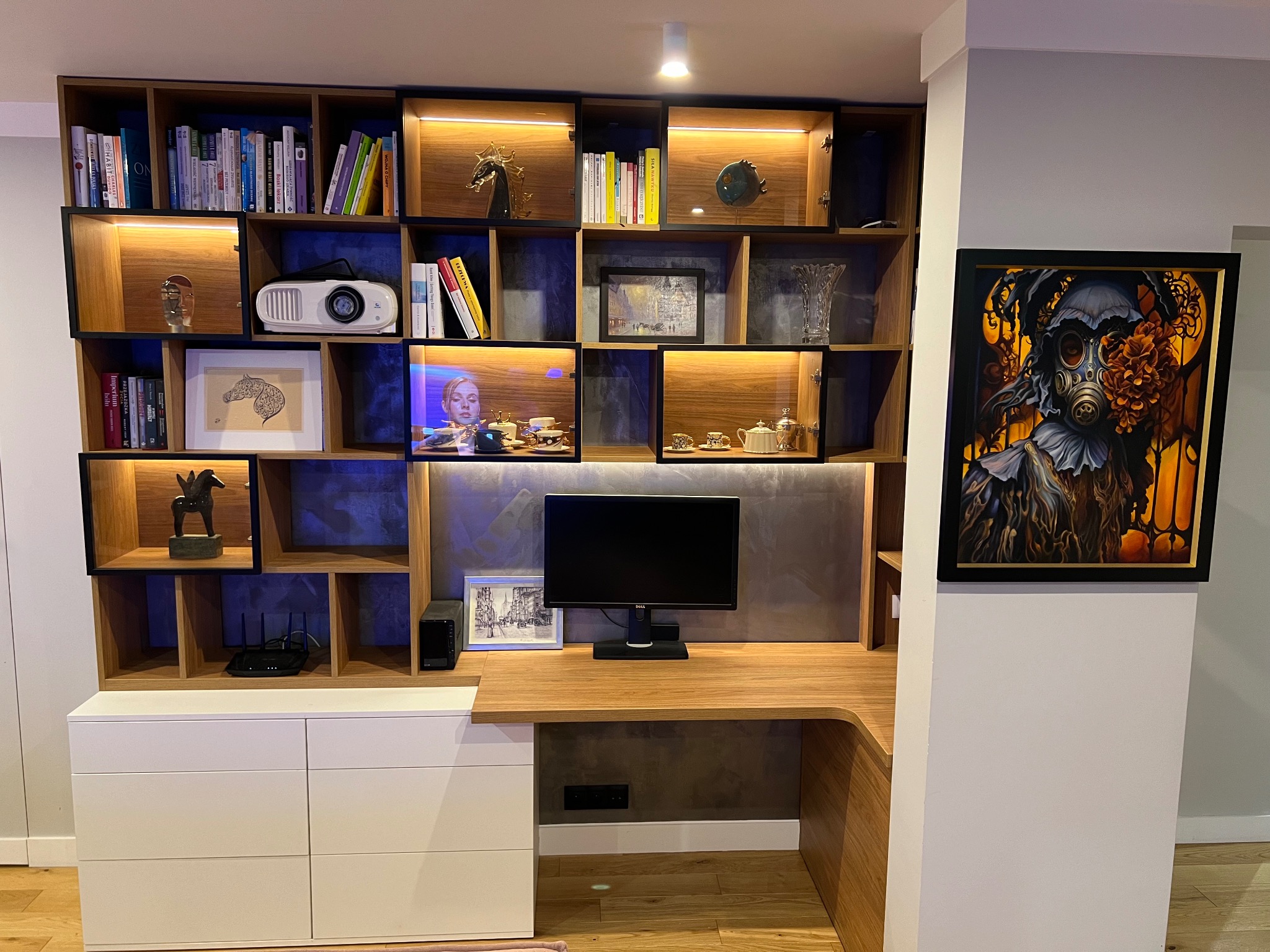 Funkcjonalna zabudowa biurowa z drewnianymi półkami, białymi szufladami i blatem. Na półkach książki, dekoracje i projektor. Na ścianie obraz w czarnej ramie.