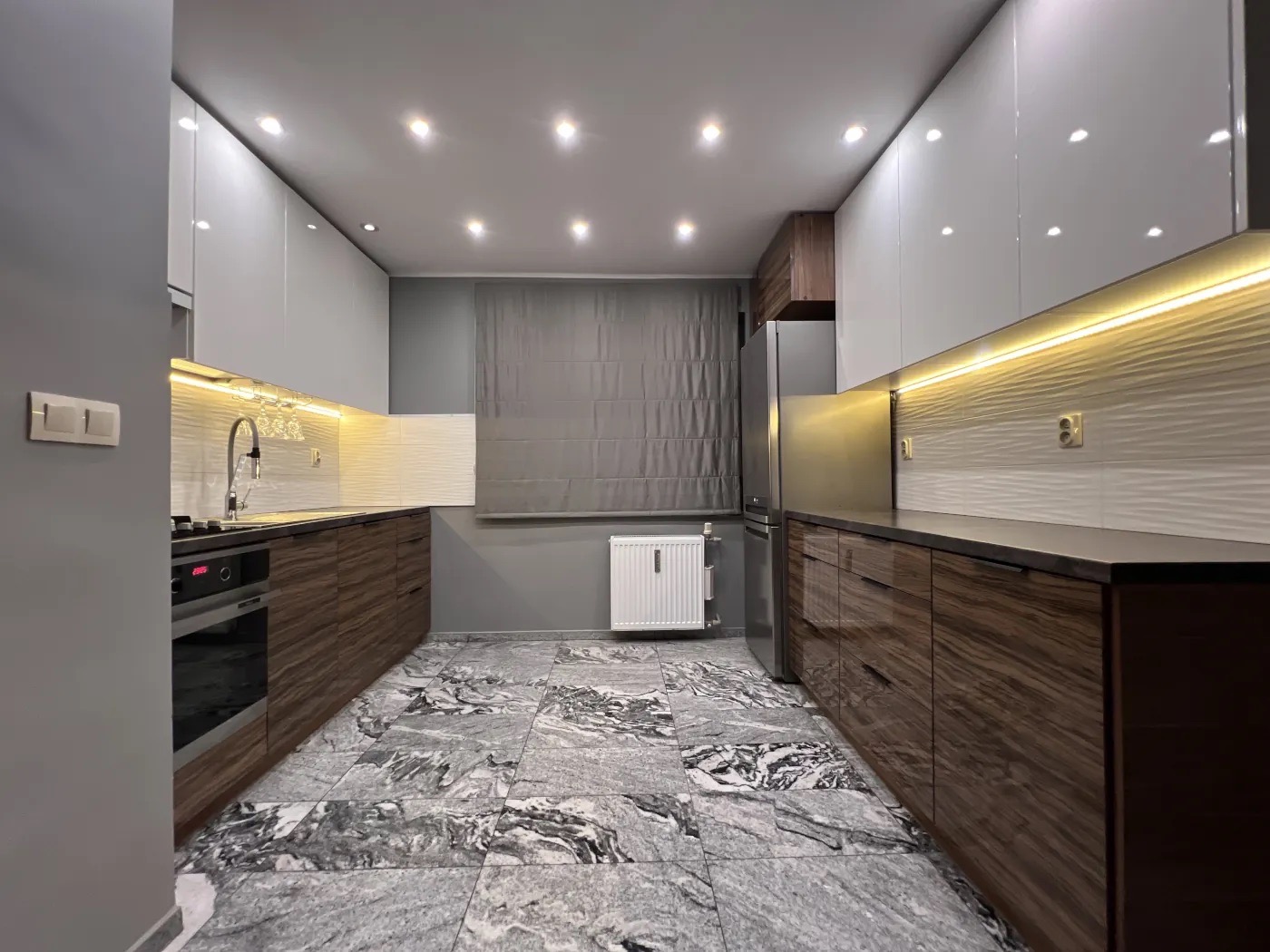 Nowoczesna kuchnia z drewnianymi szafkami dolnymi i białymi górnymi, oświetleniem LED podszafkowym i marmurową podłogą. Widok na okno z roletą rzymską.