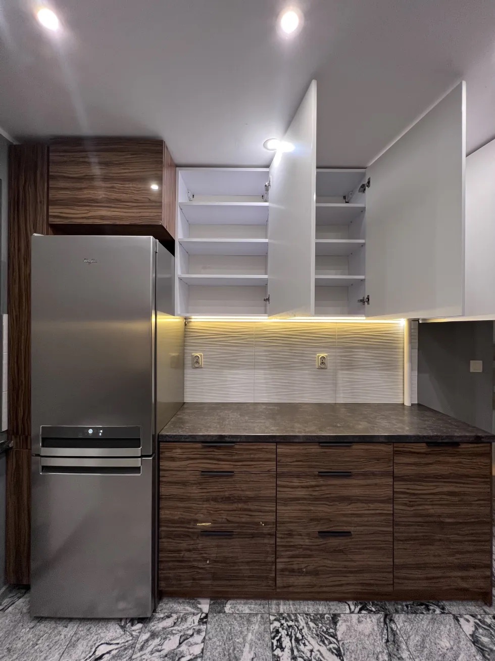 Nowoczesna kuchnia z drewnianymi szafkami dolnymi i białymi górnymi, oświetleniem LED podszafkowym i srebrną lodówką Whirlpool. Uwagę zwraca marmurowa podłoga.