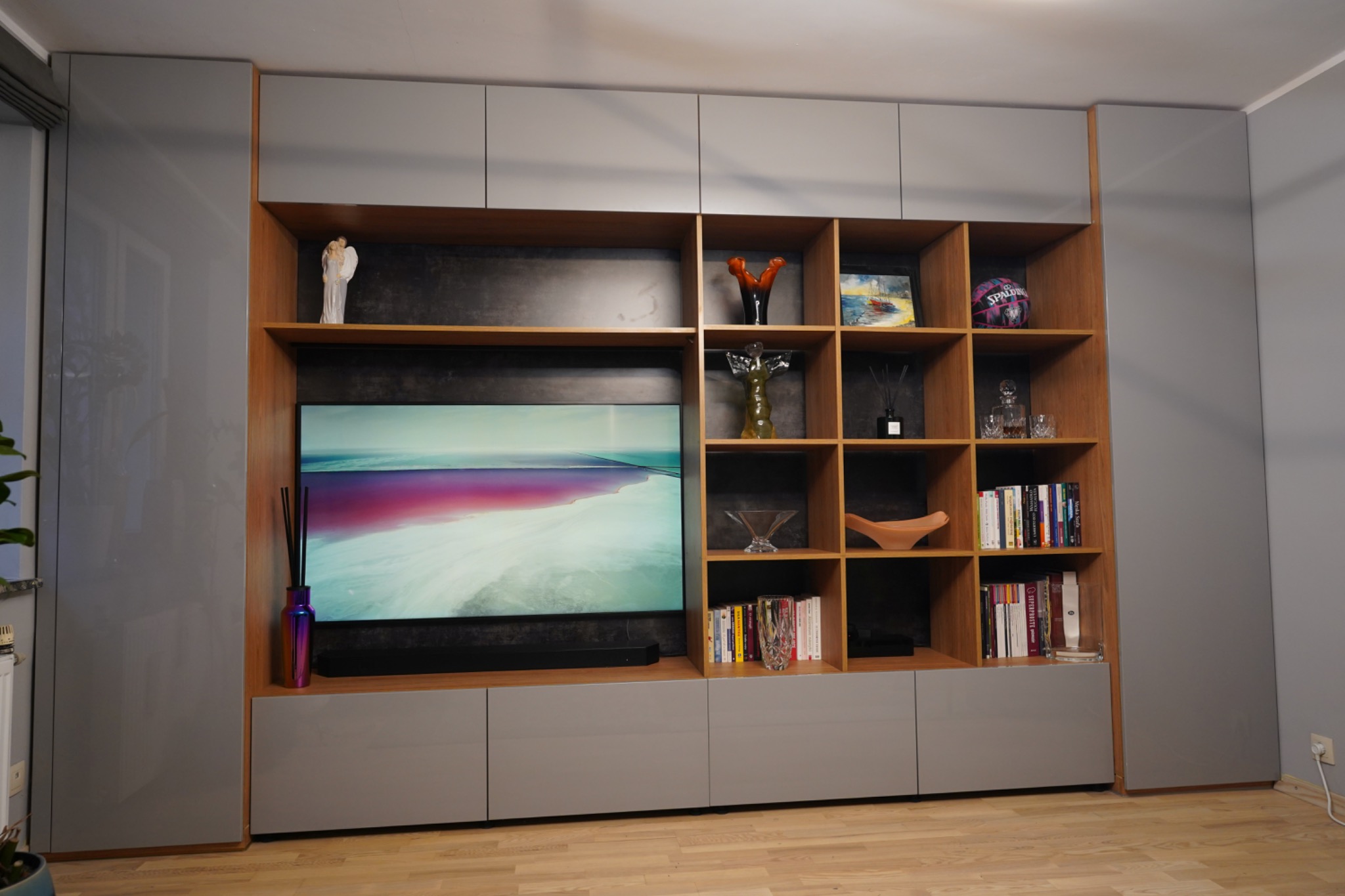 Nowoczesna zabudowa ściany z szarymi szafkami i drewnianymi półkami, na których stoją książki i dekoracje. Telewizor wkomponowany w mebel.