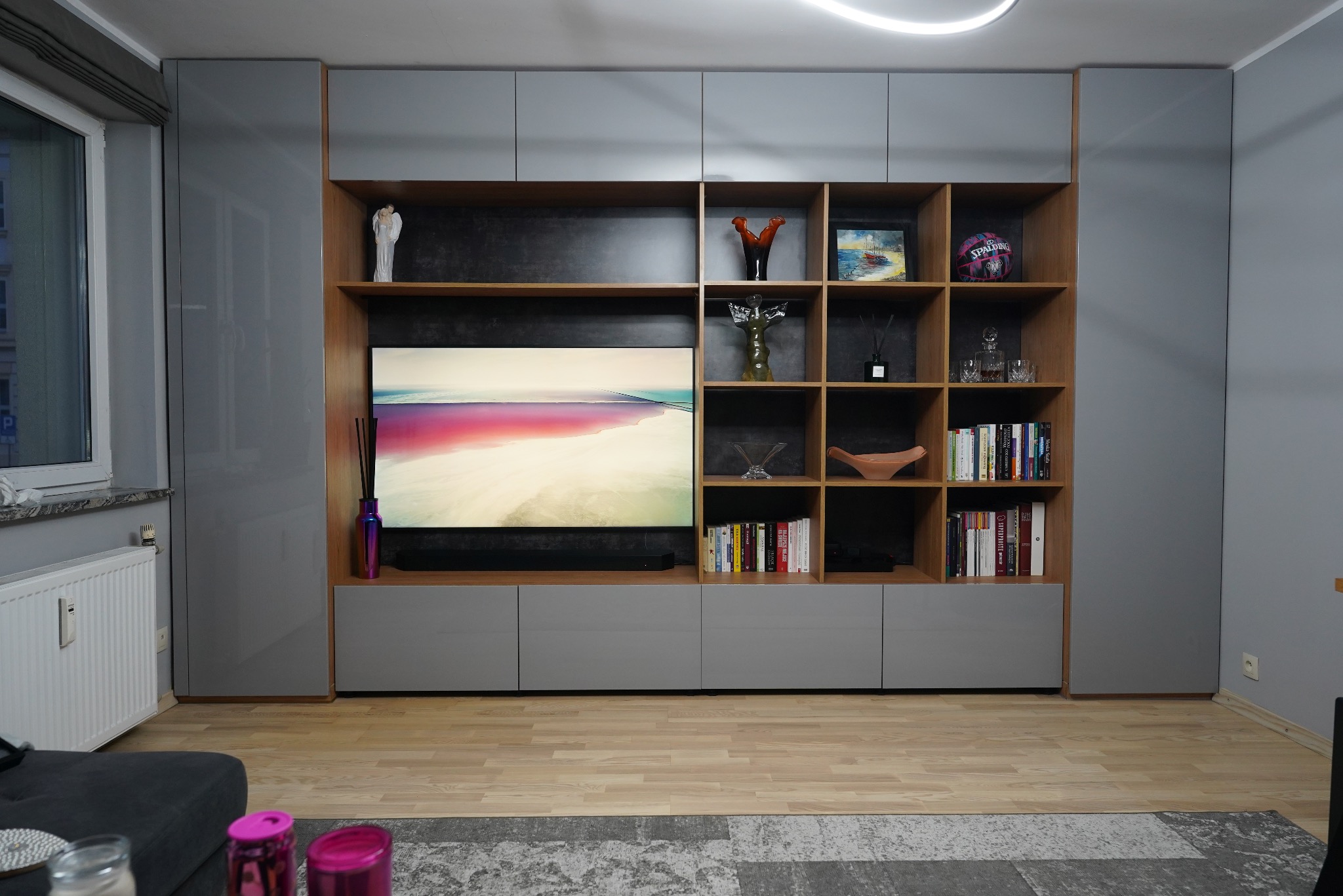 Szara szafa na wymiar z drewnianymi półkami, wnęką na telewizor i dekoracjami. Nowoczesny design, oświetlenie LED, parkiet na podłodze.