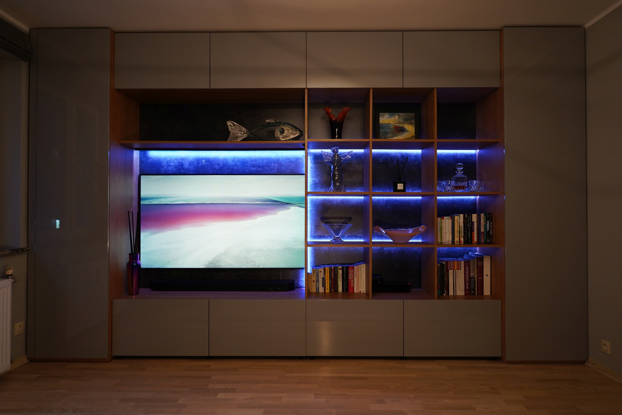 Nowoczesna szafa wnękowa z podświetleniem LED, zabudowa ściany z telewizorem. Szare fronty, drewniane półki z dekoracjami. Minimalistyczny design, ciepłe oświetlenie.