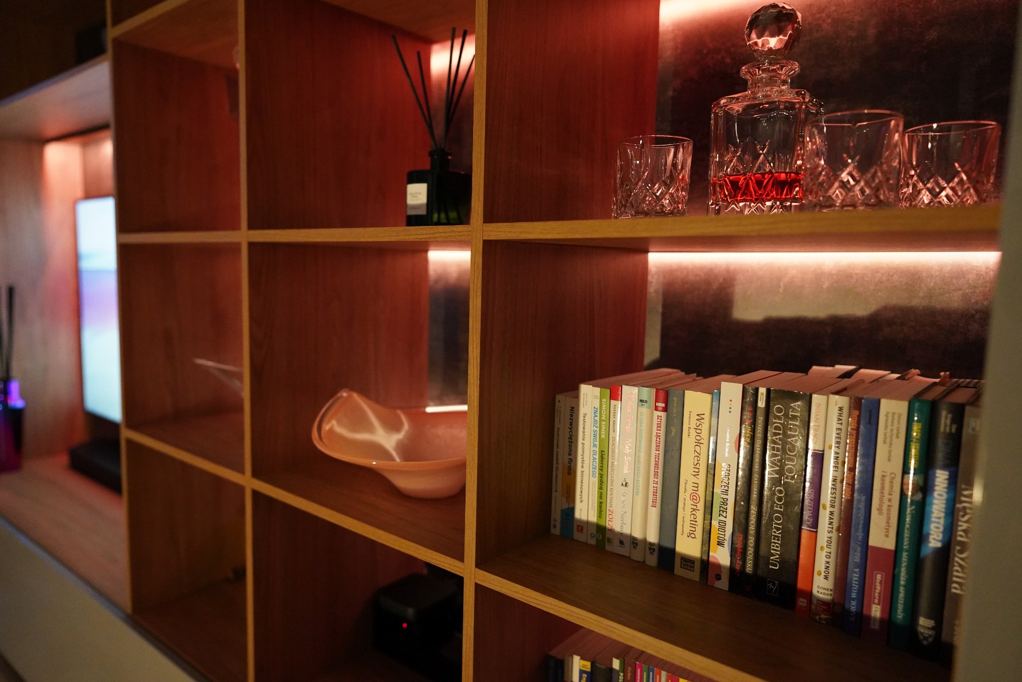 Drewniana szafka z półkami, oświetlona ciepłym światłem, eksponuje książki, naczynia i dekoracje. Widoczne detale wykończenia i aranżacji wnętrza.