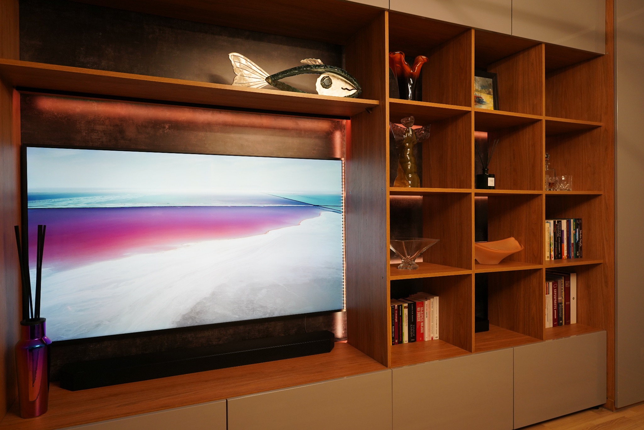 Nowoczesna szafka RTV z drewnianymi półkami i szarymi frontami. Na półkach dekoracje i książki. Telewizor z podświetleniem LED. Aranżacja wnętrza.