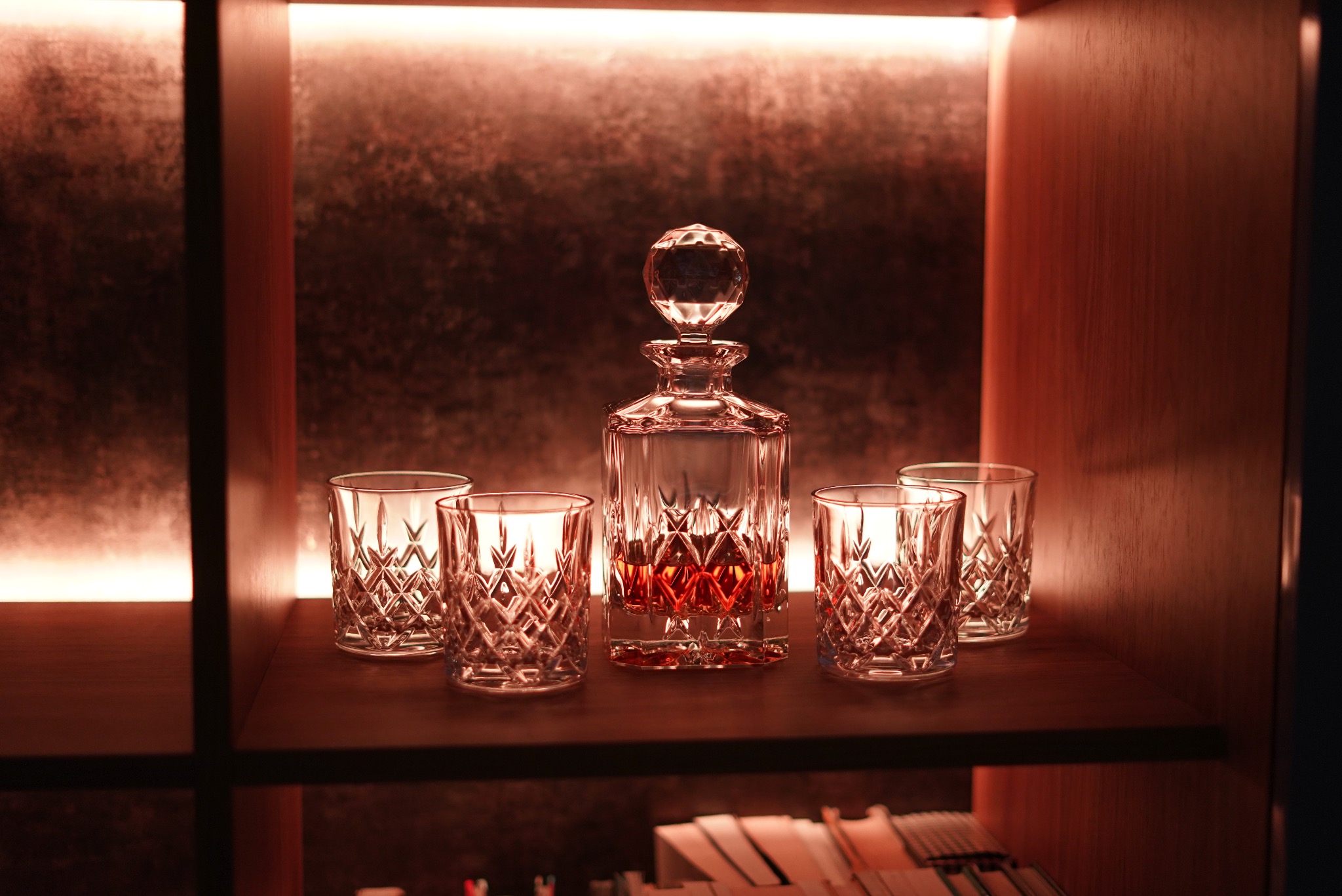 Podświetlana, drewniana półka z karafką i czterema szklankami do whisky. Elegancki barek w ciepłej tonacji.