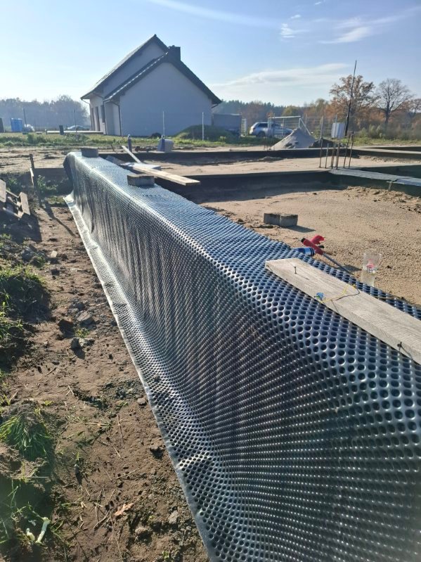 Ochrona fundamentów folią kubełkową w wykopie, zabezpieczona deskami. W tle dom jednorodzinny i plac budowy w słoneczny dzień. Detal: widoczne narzędzia.