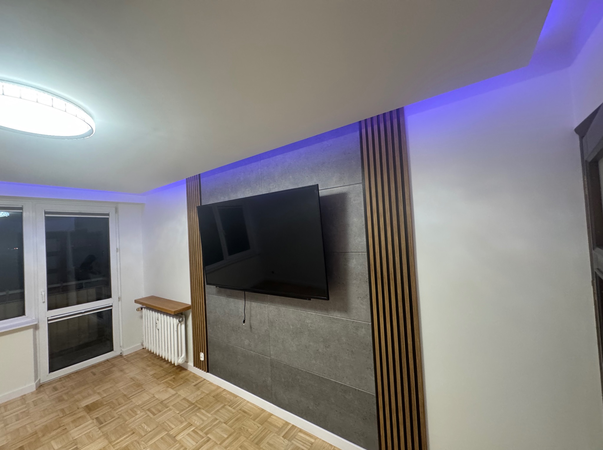 Ściana z szarych paneli i drewnianych listew, podświetlona niebieskim światłem LED, z zawieszonym telewizorem. Parkiet na podłodze, okno z lewej strony.