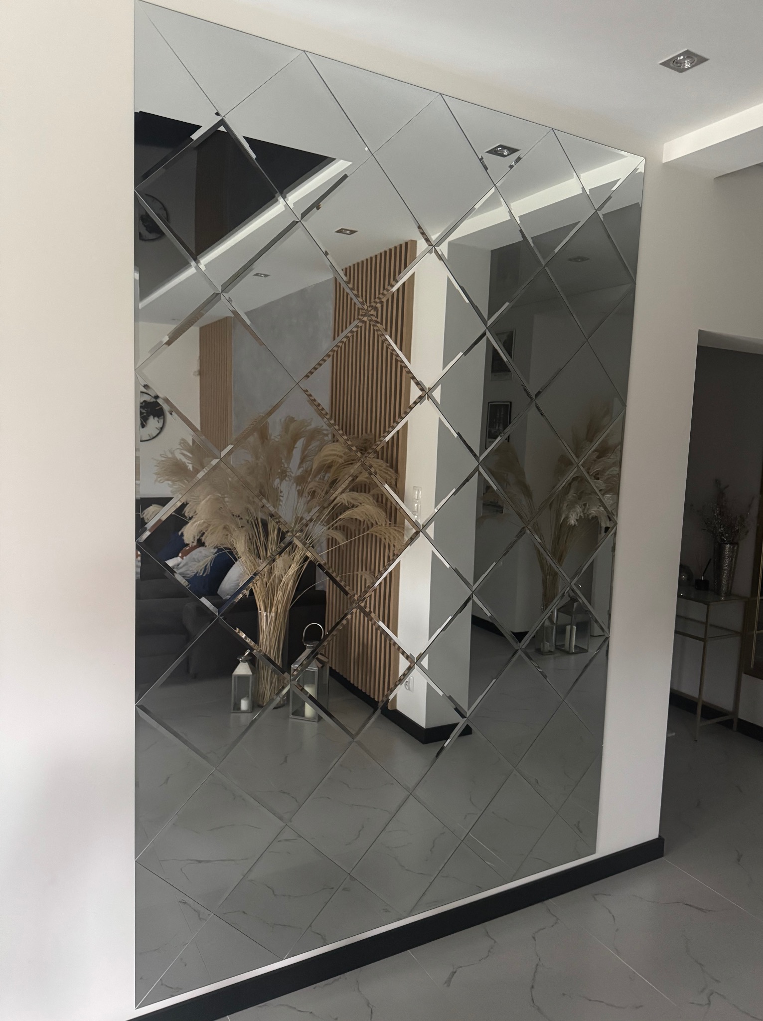 Ściana z dekoracyjnych, lustrzanych płytek w kształcie rombów, odbijająca wnętrze salonu z drewnianymi elementami i pampasową trawą w wazonie. Nowoczesny design.