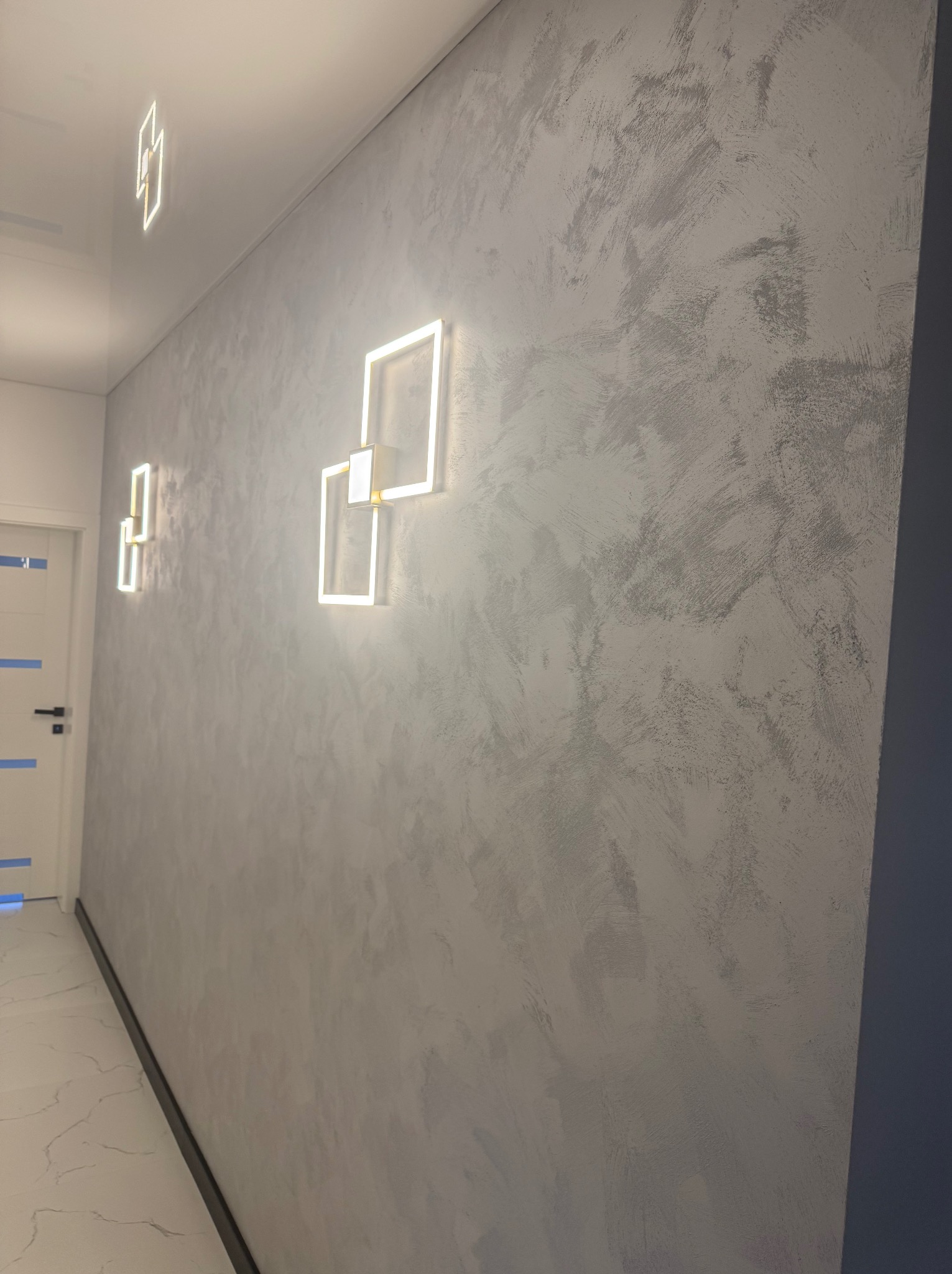 Ściana z efektem dekoracyjnym w odcieniach szarości, oświetlona nowoczesnymi, kwadratowymi lampami LED. Widoczny fragment drzwi i marmurowej podłogi.