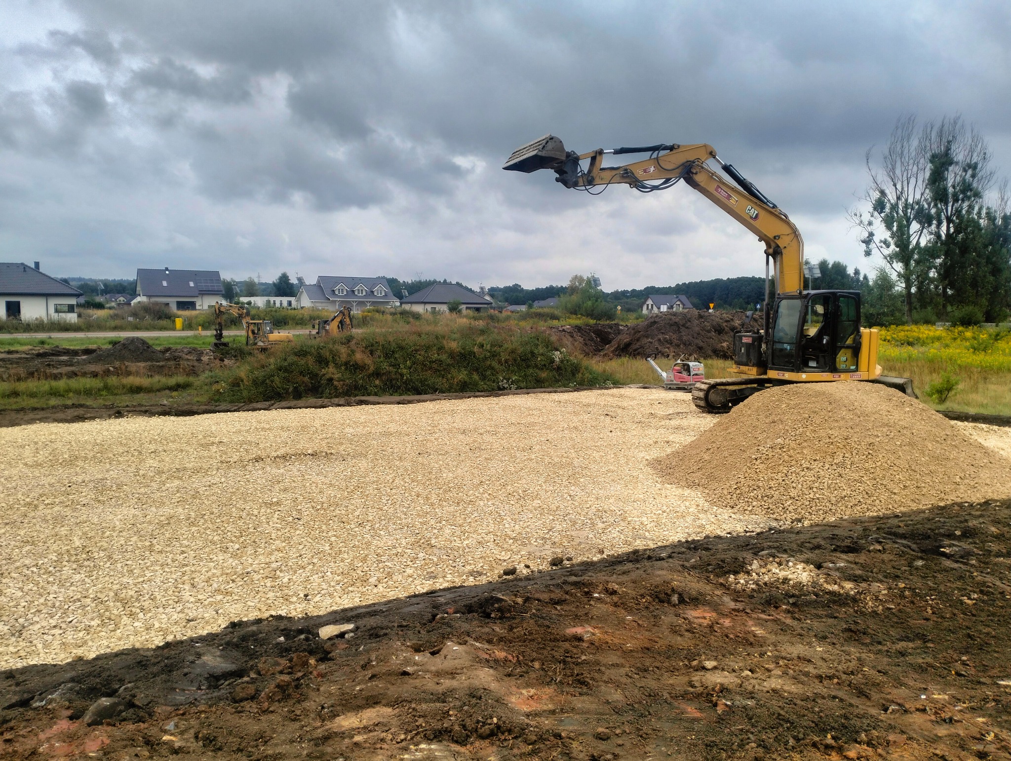 Żwirowe podłoże wyrównane koparką CAT na działce budowlanej, w tle domy jednorodzinne pod pochmurnym niebem. Przygotowanie terenu pod budowę.