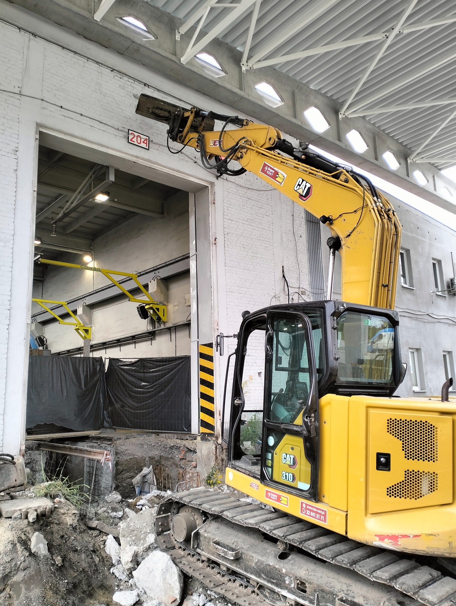 Żółta koparka CAT 310 z hydraulicznym młotem pneumatycznym przy otworze w murze budynku z numerem 204, sugerująca prace wyburzeniowe lub przygotowanie do fundamentów.