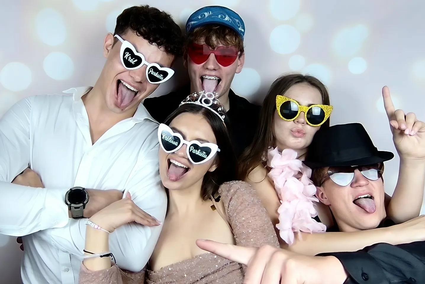 Grupa roześmianych młodych osób w okularach z napisami 'Król Parkietu' i 'Królowa Parkietu' pozuje do zdjęcia na tle jasnego tła z efektem bokeh.