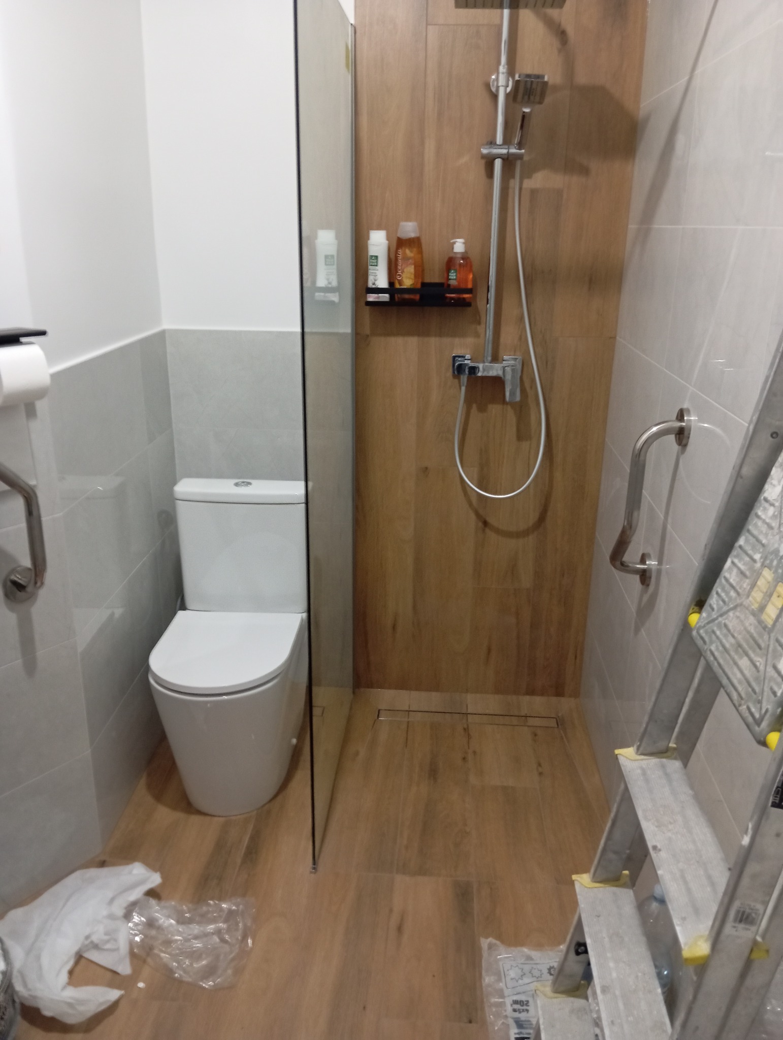 Wykończona łazienka z kabiną prysznicową typu walk-in, toaletą i drabinką. Widoczne płytki imitujące drewno i szare płytki na ścianach. Minimalistyczny design.