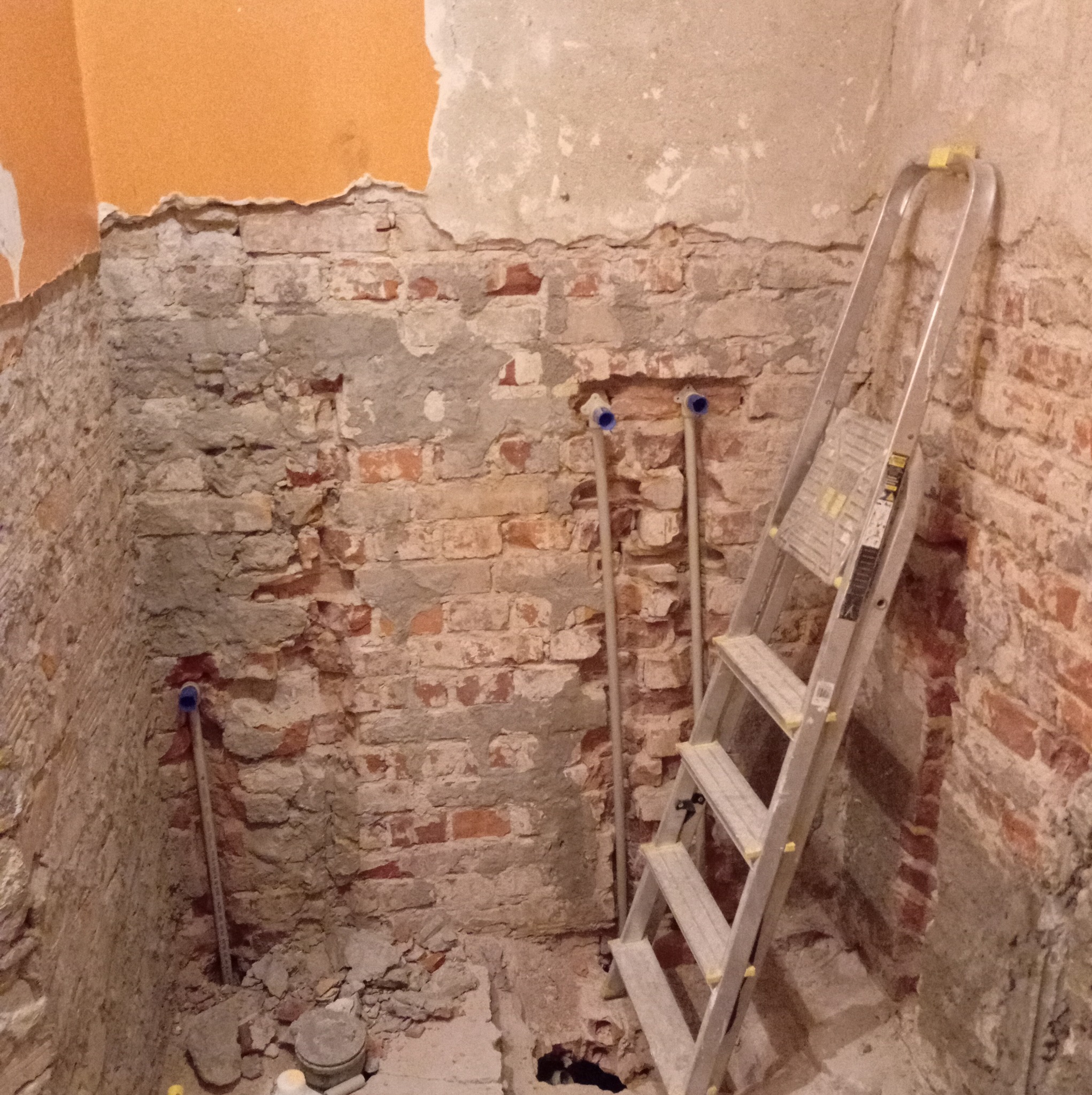 Ceglana ściana w trakcie skuwania tynku, widoczne rury instalacyjne i drabina. Fragment pomarańczowej ściany z resztkami tynku. Przygotowanie do remontu.