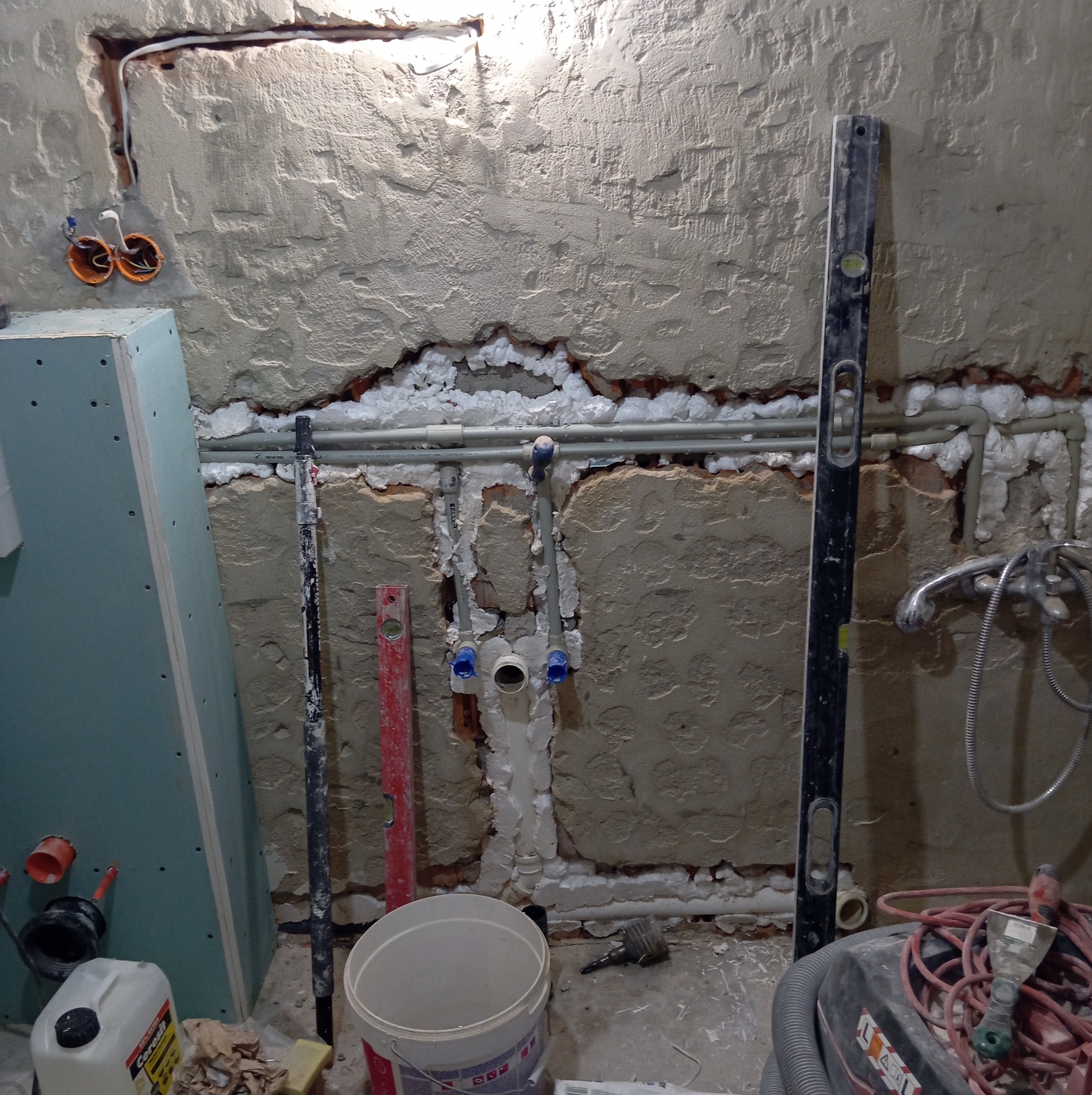 Niefotogeniczny etap prac w łazience: odkryte rury, instalacja wodna w ścianie, poziomice, płyta gipsowa i narzędzia. Widok na surową ścianę przed wykończeniem.