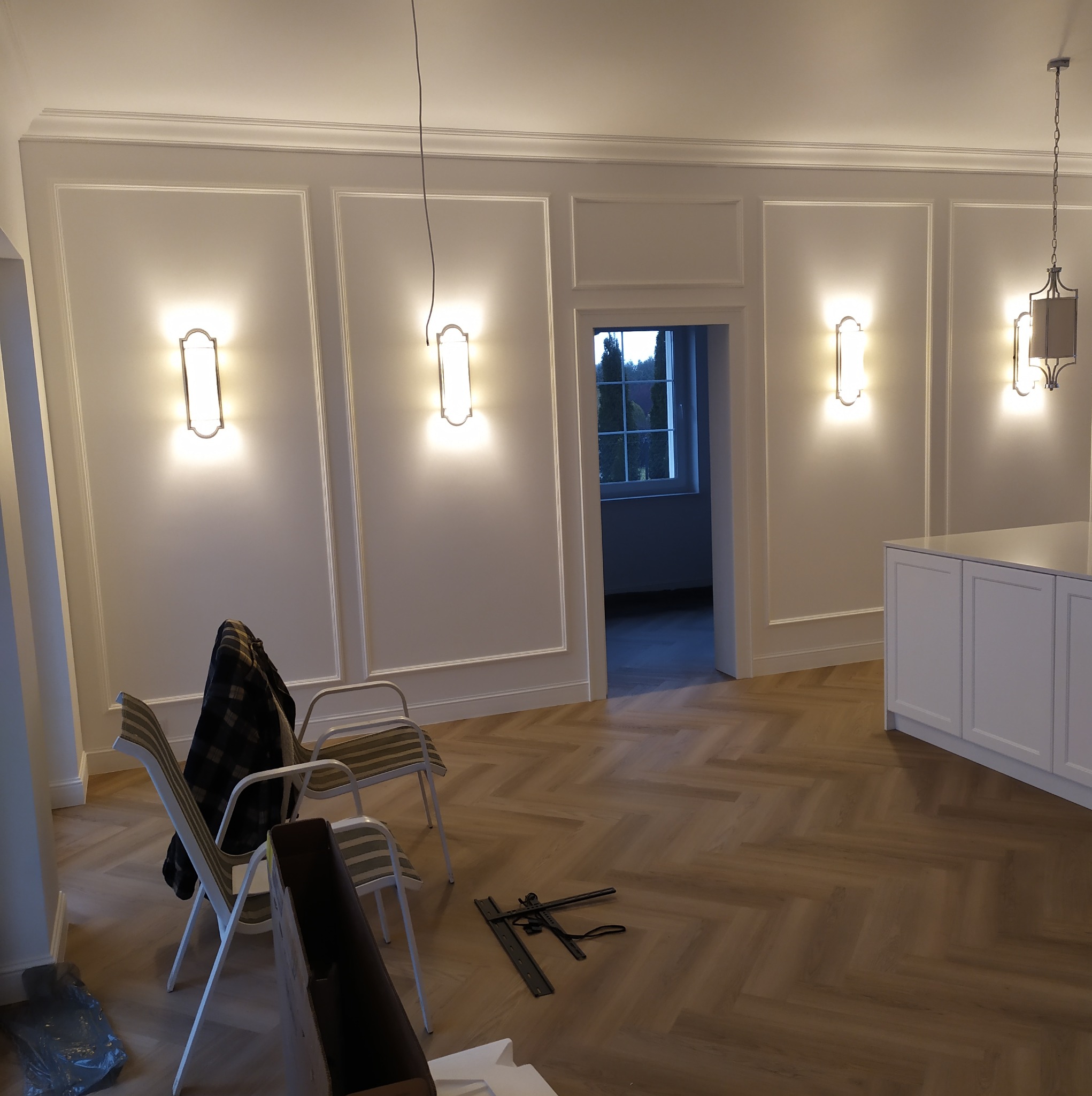 Eleganckie wykończenie wnętrza z parkietem w jodełkę, ozdobnymi panelami ściennymi i nowoczesnym oświetleniem. Widoczne krzesła i narzędzia sugerują trwające prace.