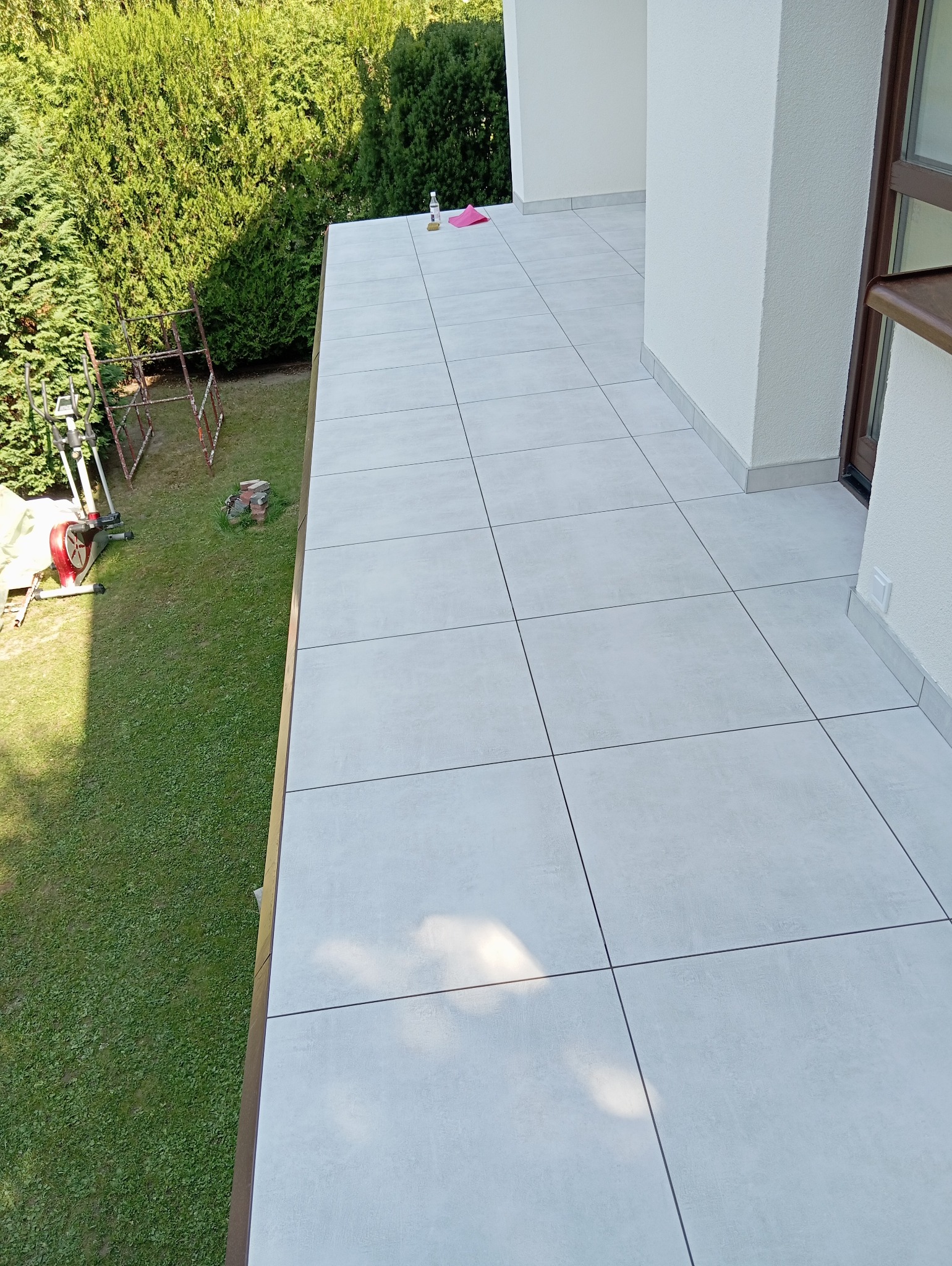 Jasnoszara, kwadratowa terakota na balkonie z widokiem na zieleń. Na płytkach leżą butelka z płynem i różowa ściereczka. W tle rusztowanie i sprzęt do ćwiczeń.