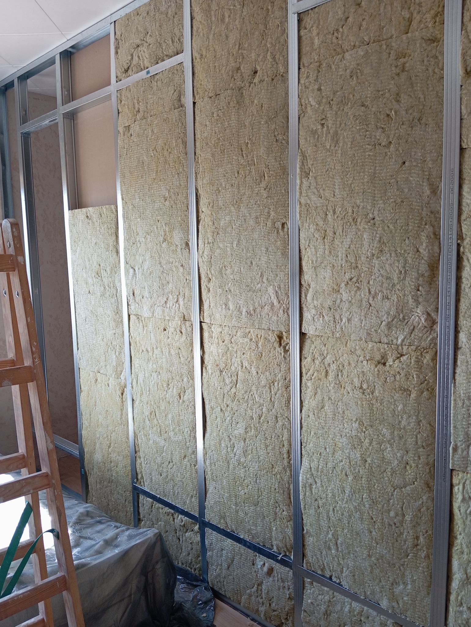 Ściana z metalowym stelażem wypełniona wełną mineralną, widoczna drabina malarska. Etap prac wykończeniowych, izolacja termiczna i akustyczna wewnątrz budynku.