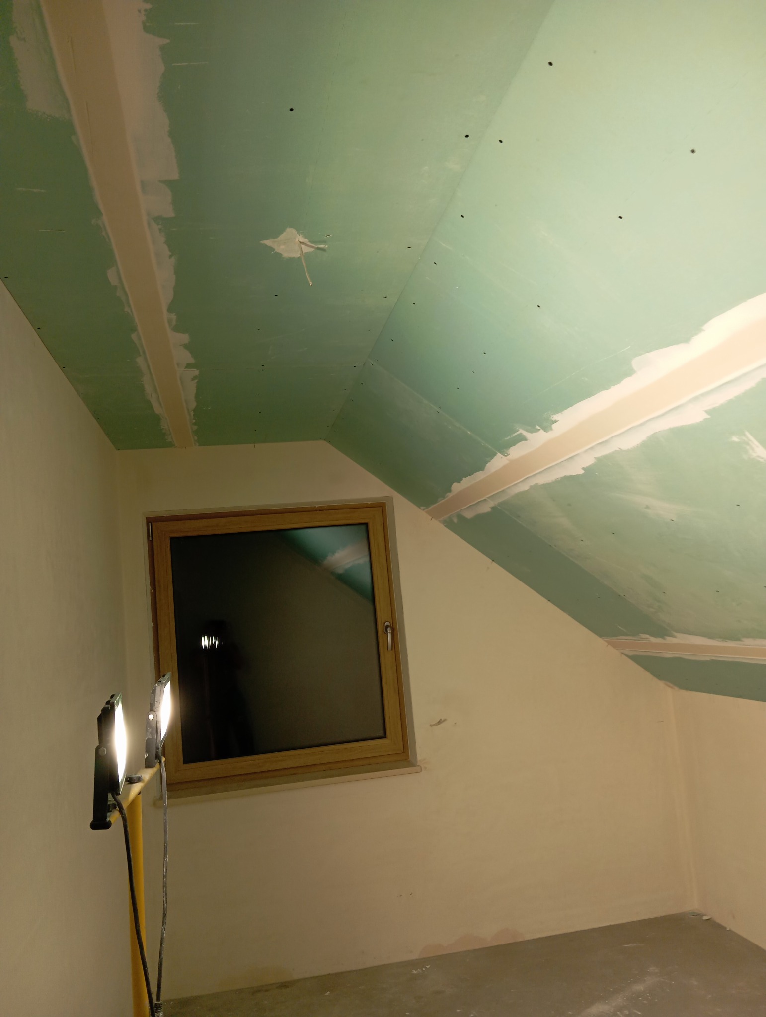 Poddasze z oknem, ściany i sufit z płyt kartonowo-gipsowych, częściowo zaszpachlowane, oświetlone lampami budowlanymi. Widoczne łączenia płyt.