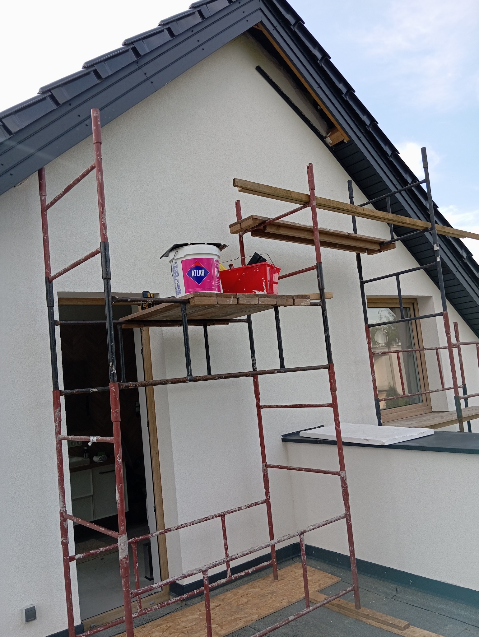 Biała elewacja domu w trakcie malowania, widoczne rusztowanie z farbą Atlas i narzędziami. Ujęcie z dołu, fragment dachu z czarną dachówką.