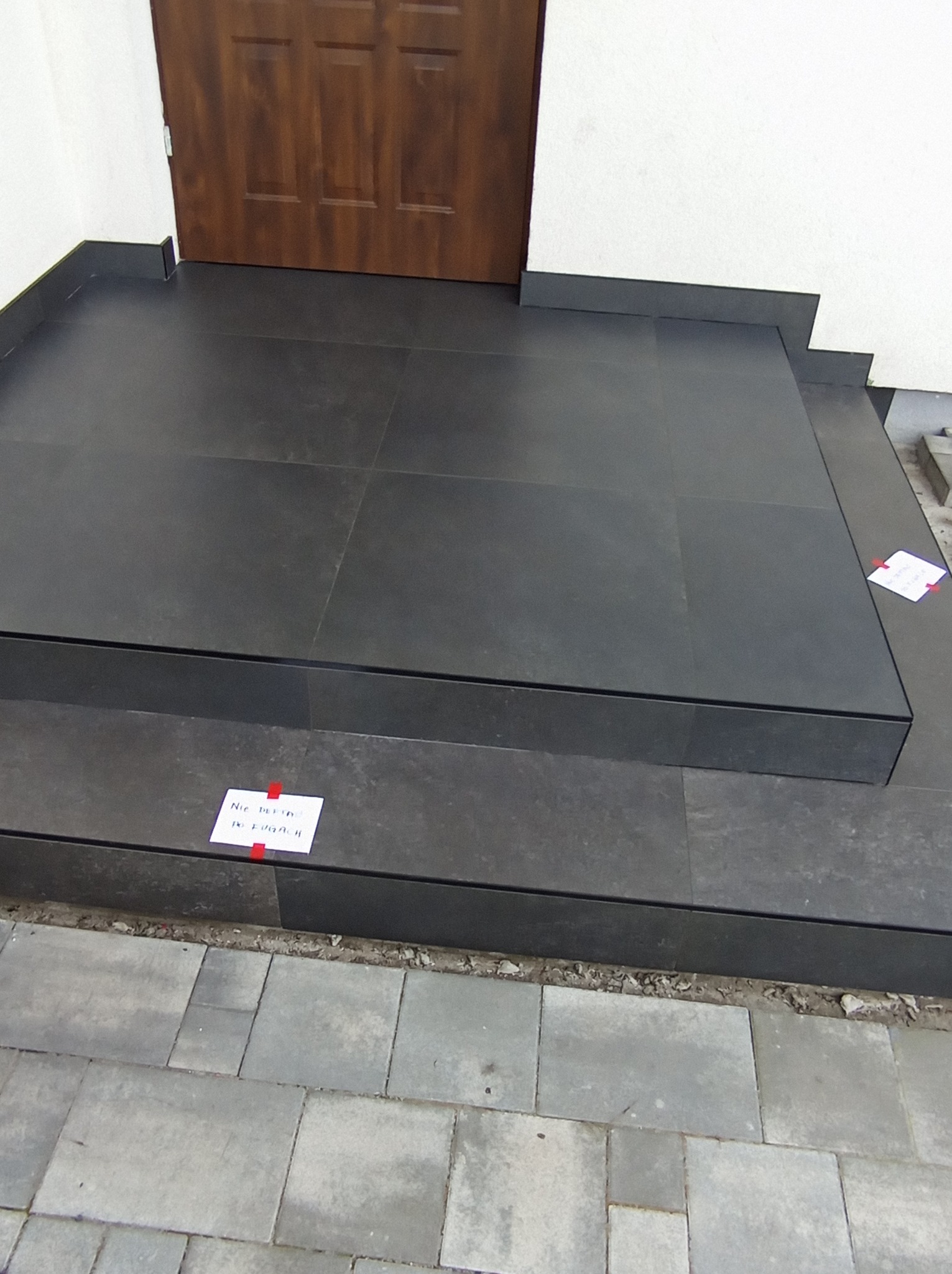 Wykończone płytkami schody wejściowe do domu z ciemnymi płytkami, widoczne detale wykończenia i fuga. Na schodach kartka z napisem.