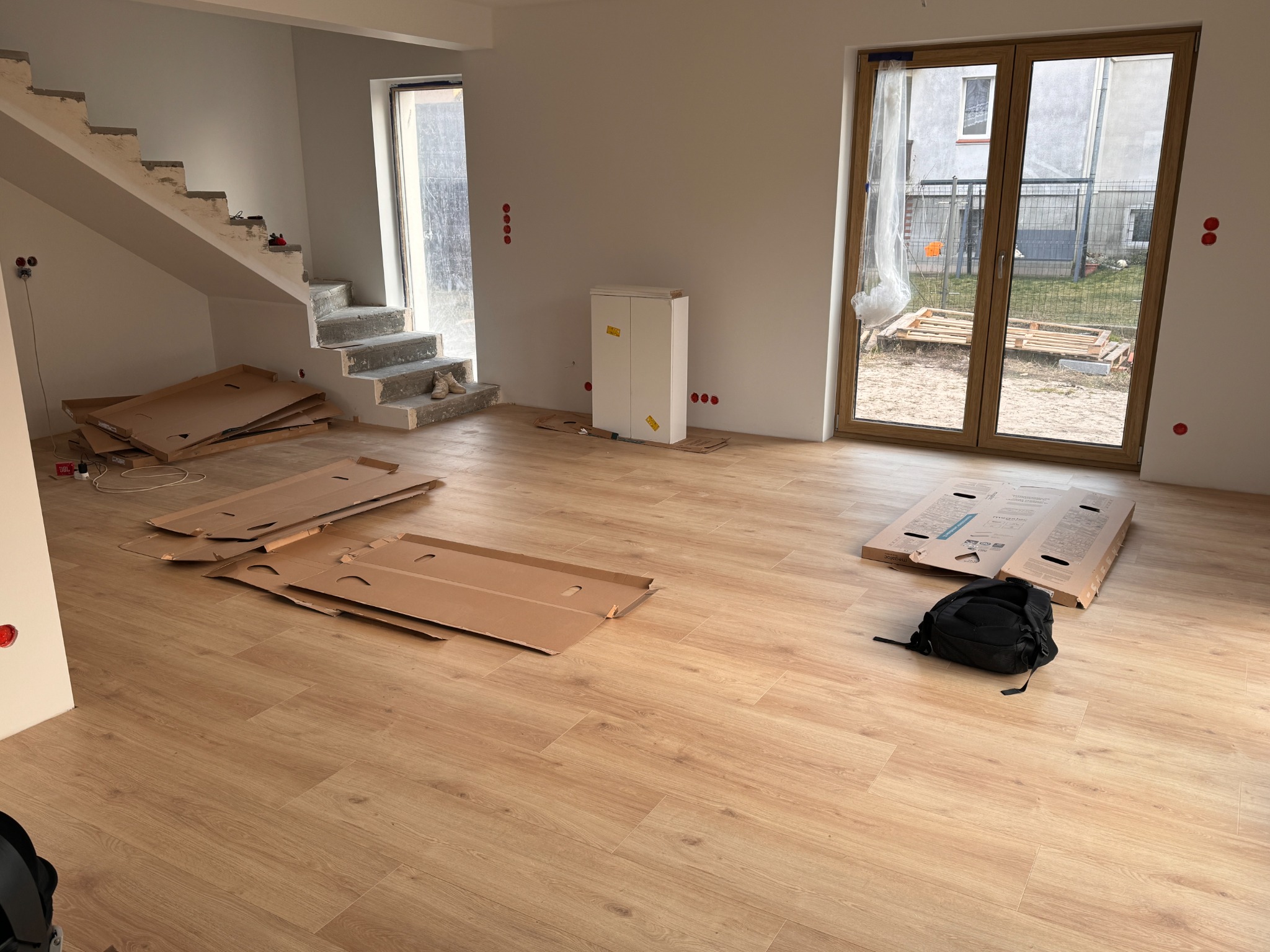 Jasne panele podłogowe ułożone w nowym domu, widoczne kartony po panelach, schody w stanie surowym, okna z widokiem na zewnątrz. Wnętrze w trakcie wykańczania.