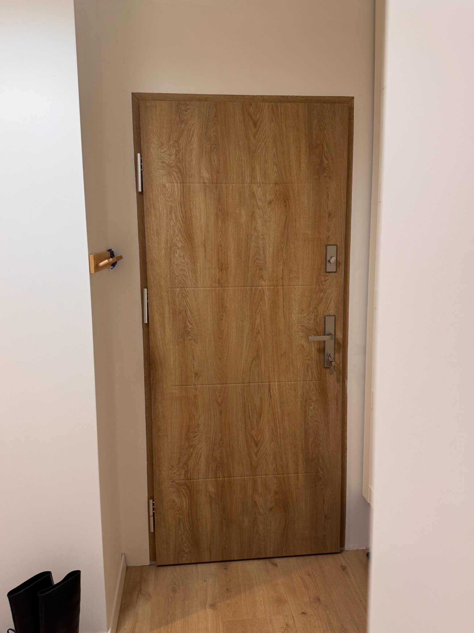 Zamknięte, drewnopodobne drzwi wejściowe z metalową klamką i zamkiem, zamontowane w białej ścianie. Widoczny fragment podłogi i wieszak na ścianie.