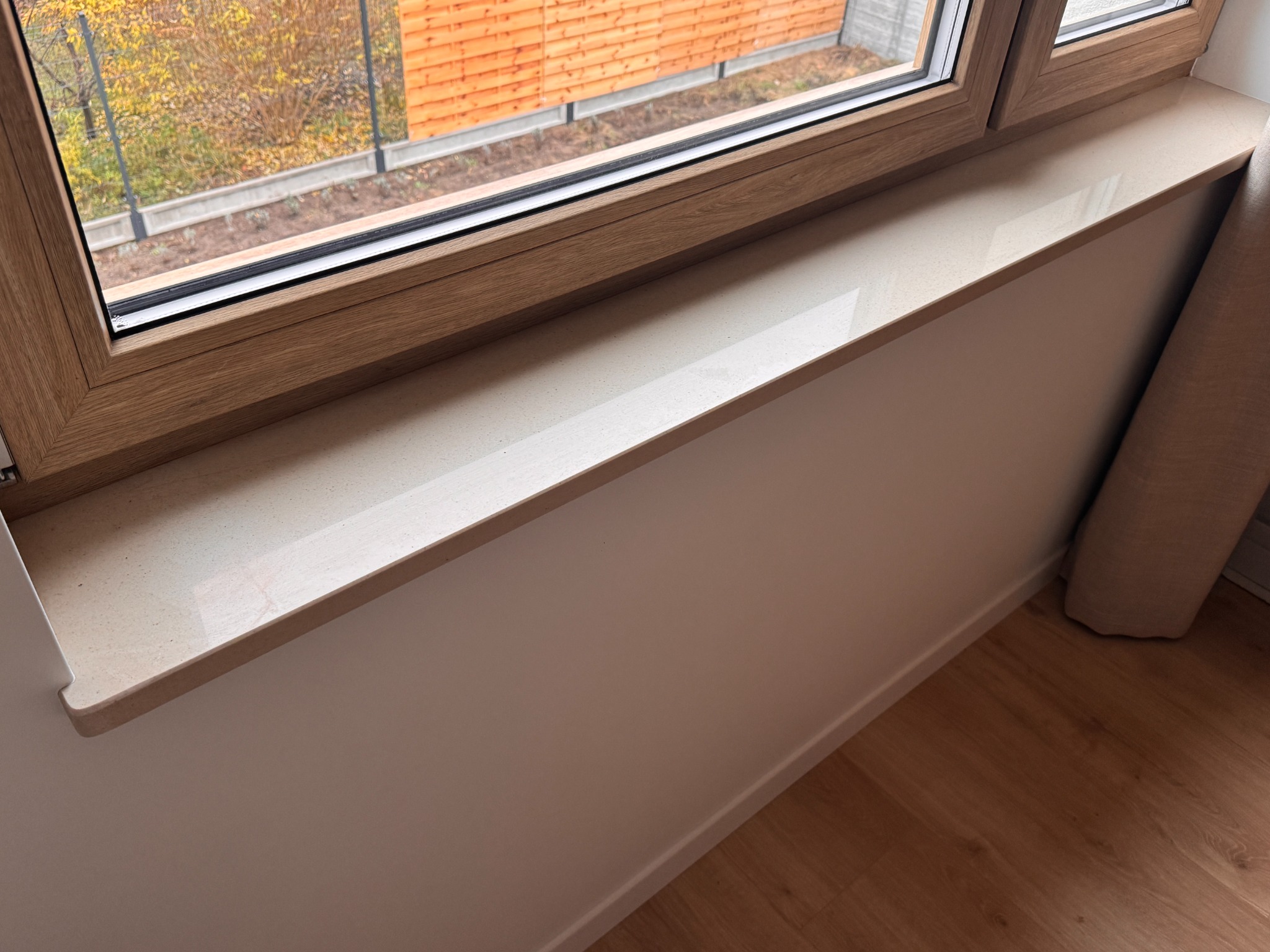 Jasny parapet z konglomeratu kwarcowego, zamontowany w oknie z drewnianą ramą. Widok na zewnątrz, na drzewa i ogrodzenie. Podłoga drewniana.