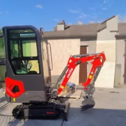 Czerwono-czarna mini koparka gąsienicowa IHIte na betonowym podłożu, z otwartą kabiną i widocznym ramieniem kopiącym, na tle budynku z drewnianą elewacją.