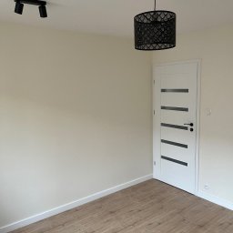 Firma Ogólnobudowlana Damian Jurak - Jasne wnętrze po remoncie: panele podłogowe, kremowe ściany, białe drzwi z czarnymi akcentami, lampa sufitowa i reflektor. Minimalistyczny design.