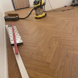 Firma Ogólnobudowlana Damian Jurak - Ułożona podłoga z paneli w jodełkę, poziomica, listwa przypodłogowa, odkurzacz przemysłowy. Widok na fragment pomieszczenia w trakcie prac wykończeniowych.