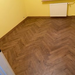 Firma Ogólnobudowlana Damian Jurak - Ukończone układanie paneli w jodełkę w pokoju z żółtymi ścianami. Widoczny grzejnik i okno. Nowa podłoga z drewnopodobnych paneli. Listwy przypodłogowe zamontowane.
