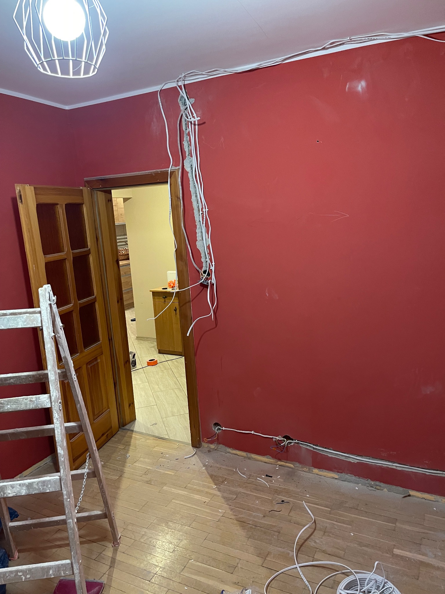 Czerwona ściana w trakcie remontu z widoczną instalacją elektryczną, drewnianą drabiną i fragmentem parkietu. Widoczne przewody i otwarta przestrzeń do innego pomieszczenia.