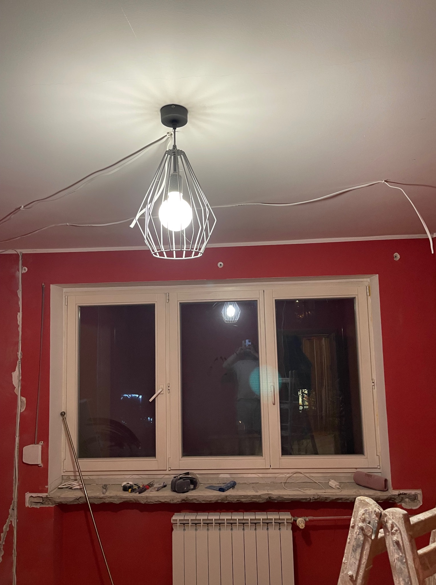 Czerwona ściana w trakcie remontu z oknem, na parapecie narzędzia, drabina i lampa wisząca. Widoczne kable na suficie i ślady po pracach wykończeniowych.