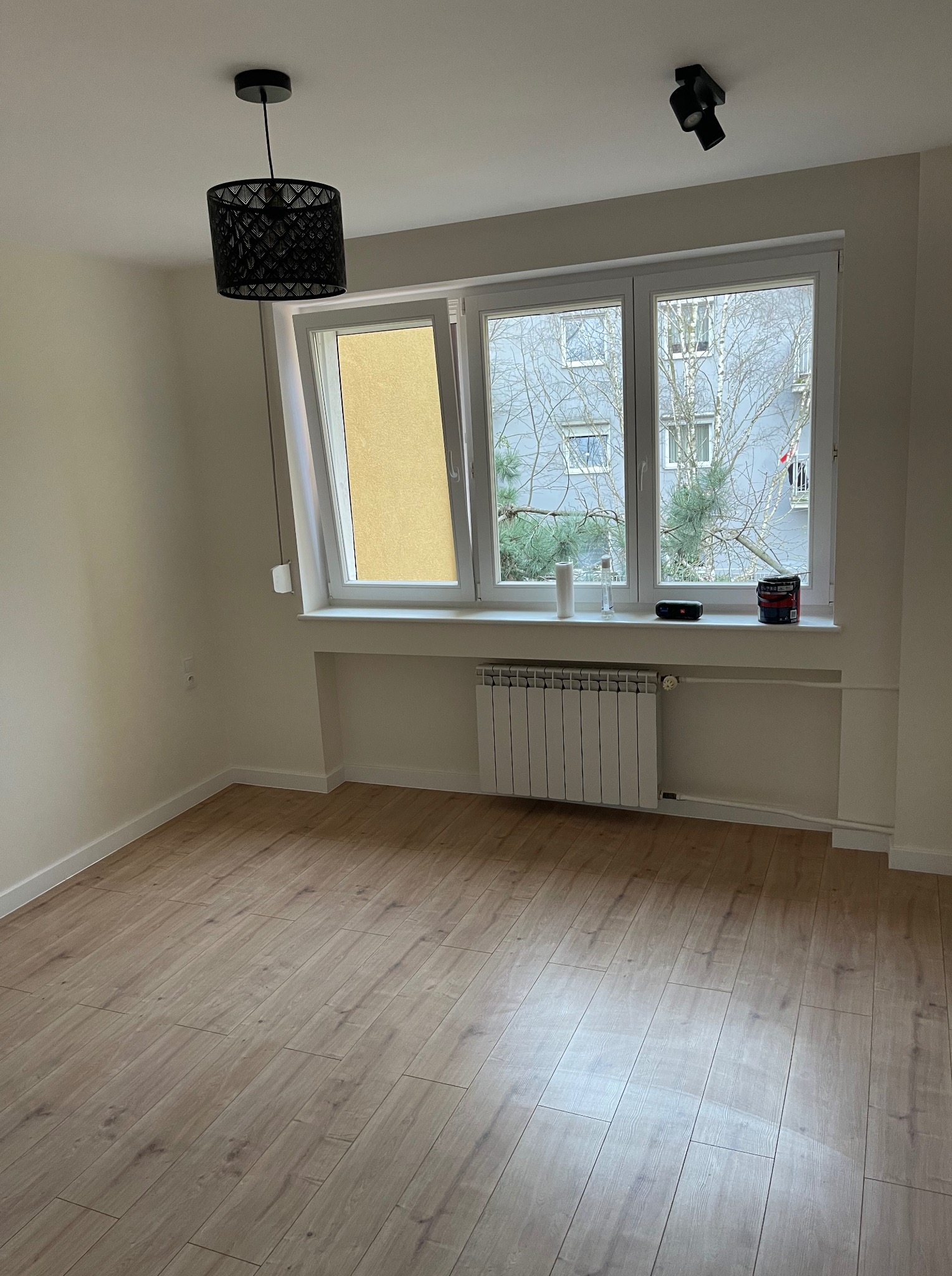 Jasne panele podłogowe w pustym pokoju z oknem, przez które widać drzewa. Na parapecie puszka farby i głośnik. Czarna lampa sufitowa i reflektory.